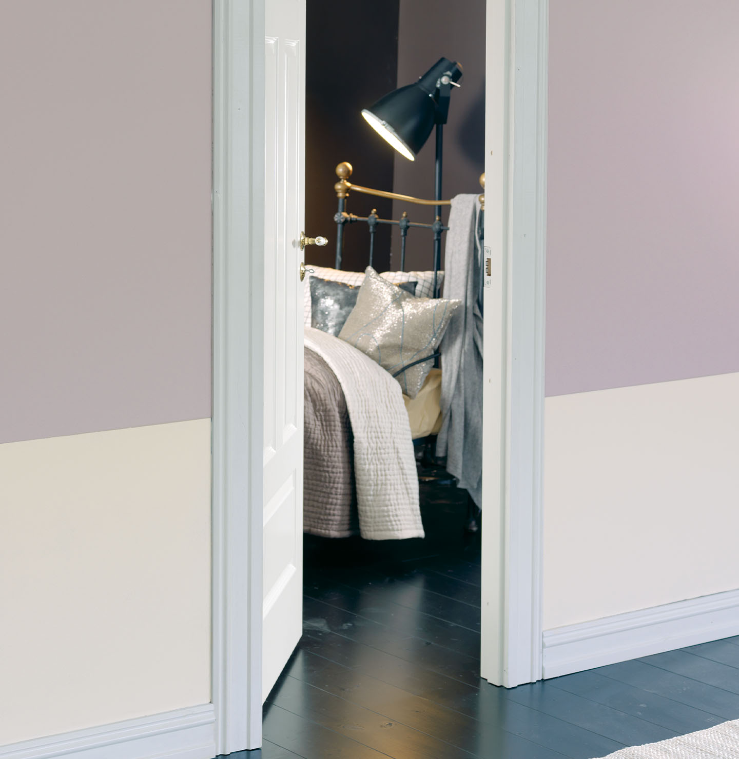 Puerta abierta que revela un dormitorio acogedor con una cama de estructura metálica, visto desde un pasillo con pared violeta suave pintada con Jotun 2525 Pilates.