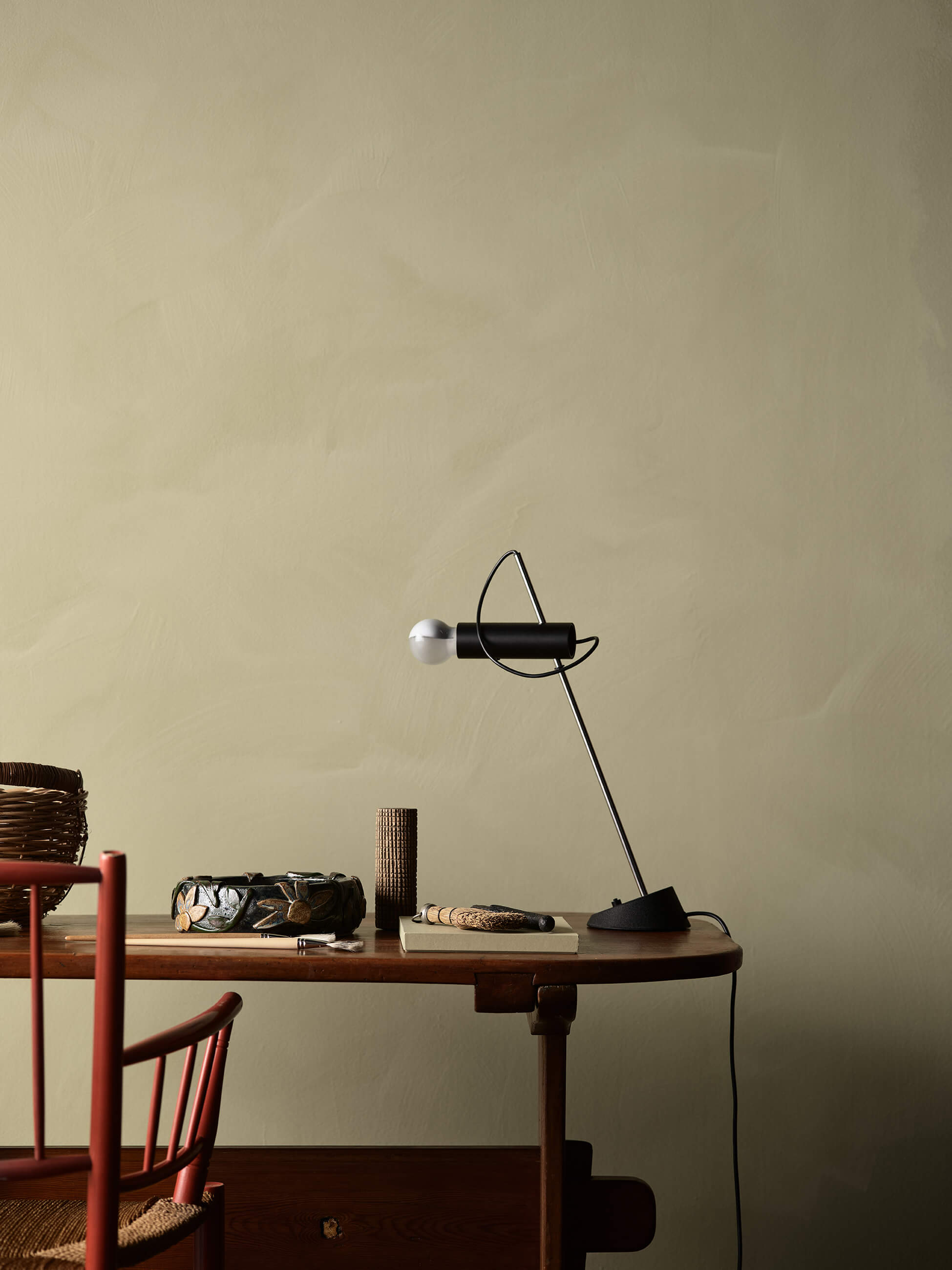 Et hjemmekontor med kalkmalte olivengrønne vegger i Jotun 0290 Biri i LADY Minerals, et skrivebord i tre med en svart bordlampe og rustikke gjenstander.