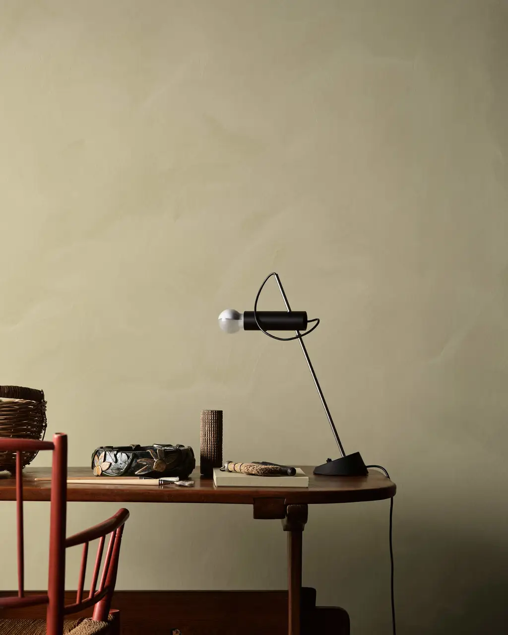 Et hjemmekontor med kalkmalede olivengrønne vægge i Jotun 0290 Biri fra LADY Minerals, et skrivebord i træ med en sort bordlampe samt rustikke genstande, der skaber en varm, naturlig og inspirerende atmosfære.