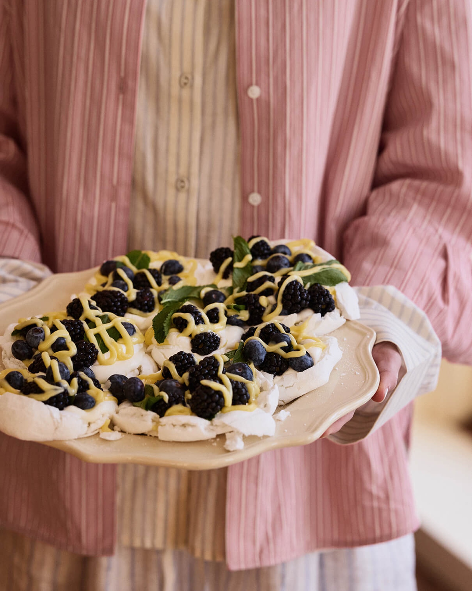 Et nærbilde av en person i en rosa-stripete skjorte som holder et fat med marengsdesserter toppet med blåbær, bjørnebær og gul saus.