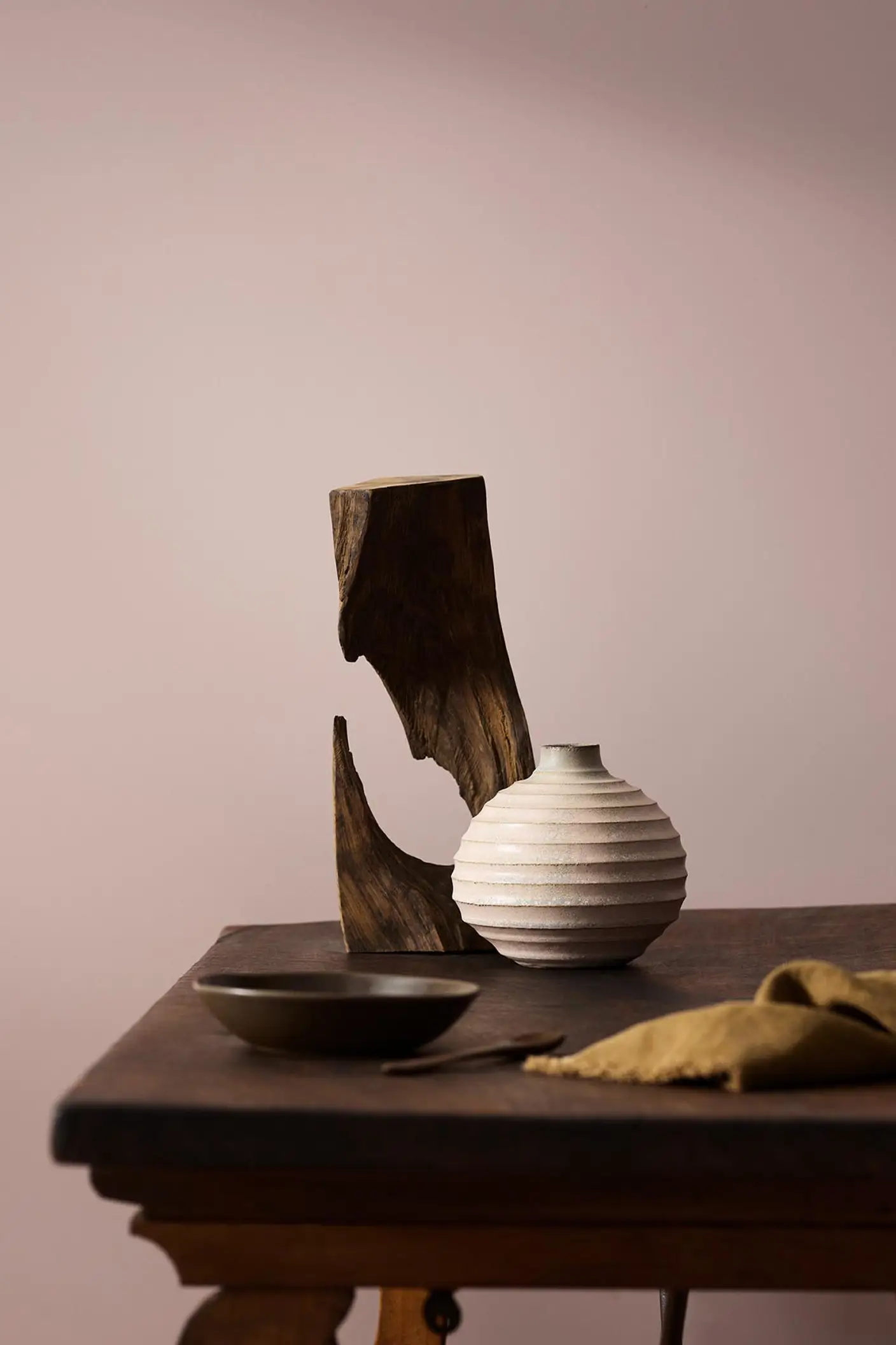 Wooden table with a ceramic vase, sculpted wood, and a dark bowl against a muted pink background painted with the colour 20054 Silky Pink from Jotun.