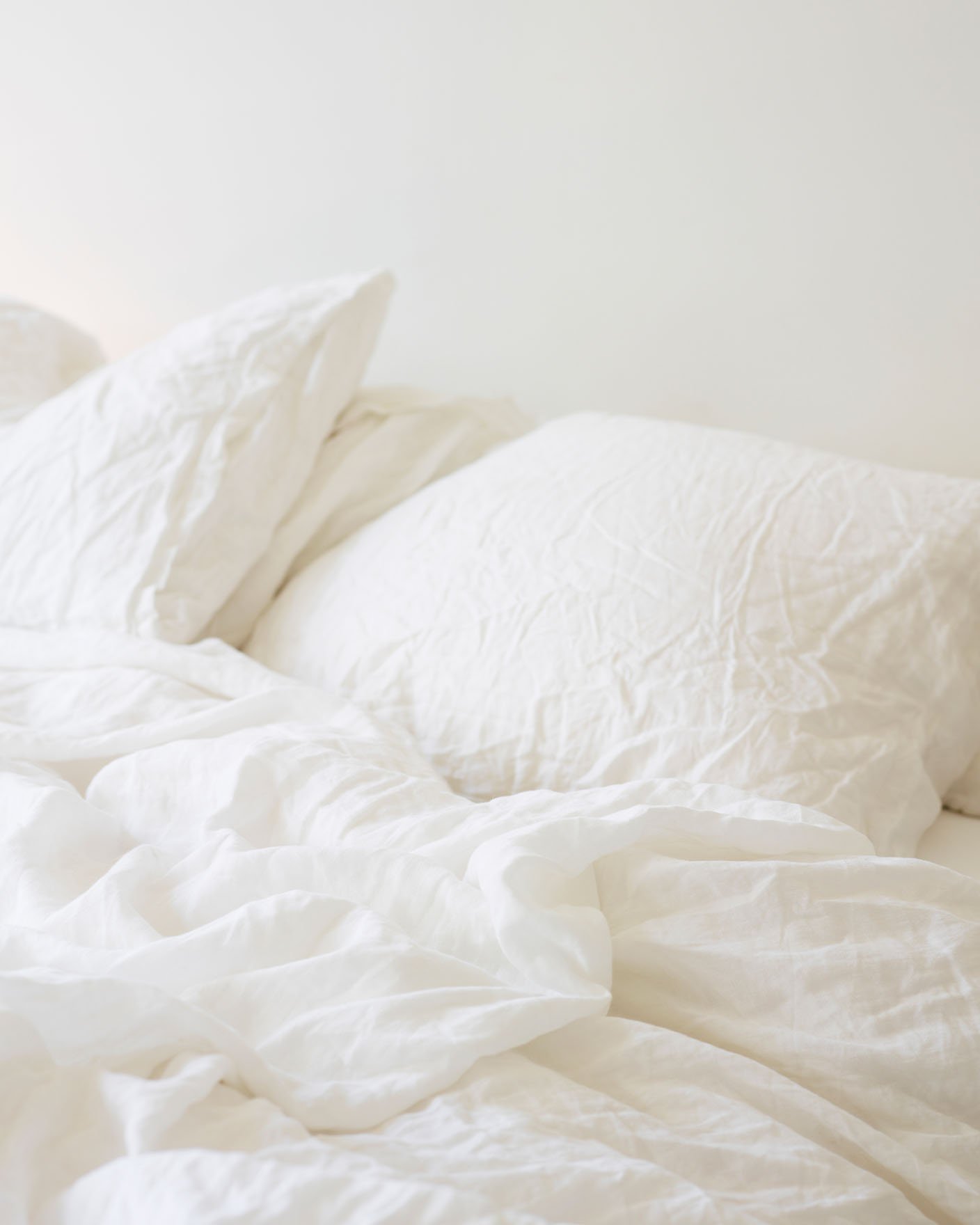 Obäddad säng med mjukt, skrynkligt vitt linnelakan och kuddar mot en varm, vitmålad vägg i Jotun 1622 Reflektion.