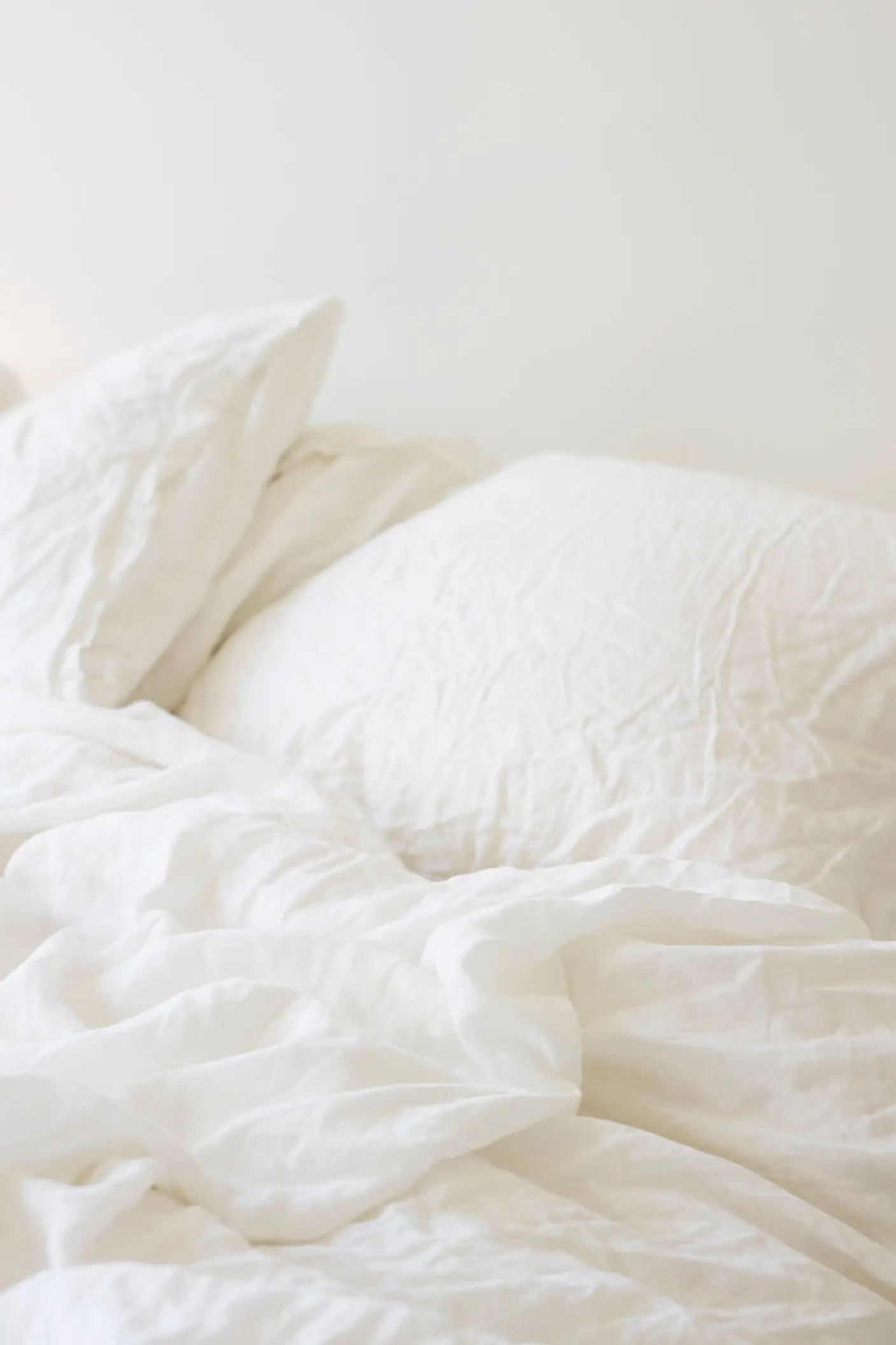 Obäddad säng med mjukt, skrynkligt vitt linnelakan och kuddar mot en varm, vitmålad vägg i Jotun 1622 Reflektion.