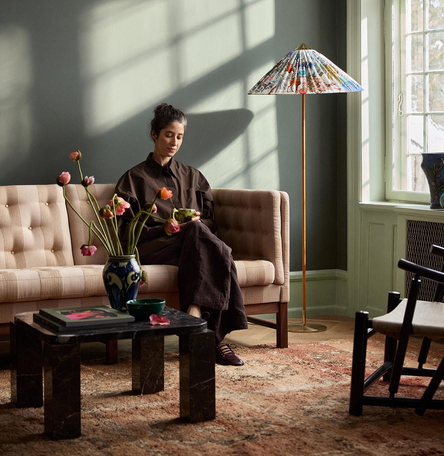 Woman sitting in a sofa with sunlight streaming through a window. The background wall is painted with the dark, muted green colour 7613 Northern Mystic from Jotun.