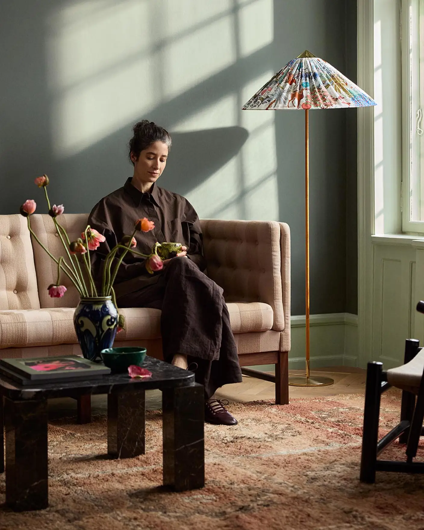 Woman sitting in a sofa with sunlight streaming through a window. The background wall is painted with the dark, muted green colour 7613 Northern Mystic from Jotun.