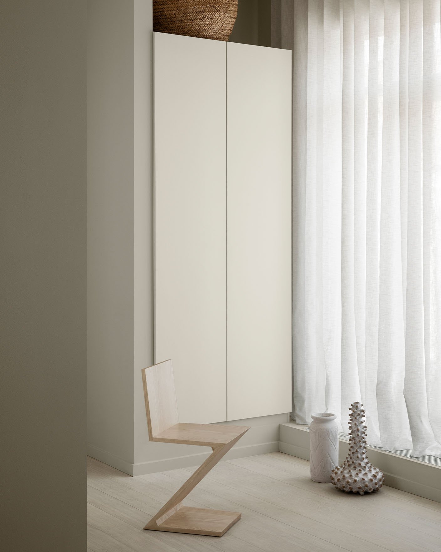 Minimalist interior with a geometric wooden chair, a cabinetry painted in Jotun 1376 Mist, and sheer white curtains. Wall in front painted in 12180 Present, a muted beige from Jotun.