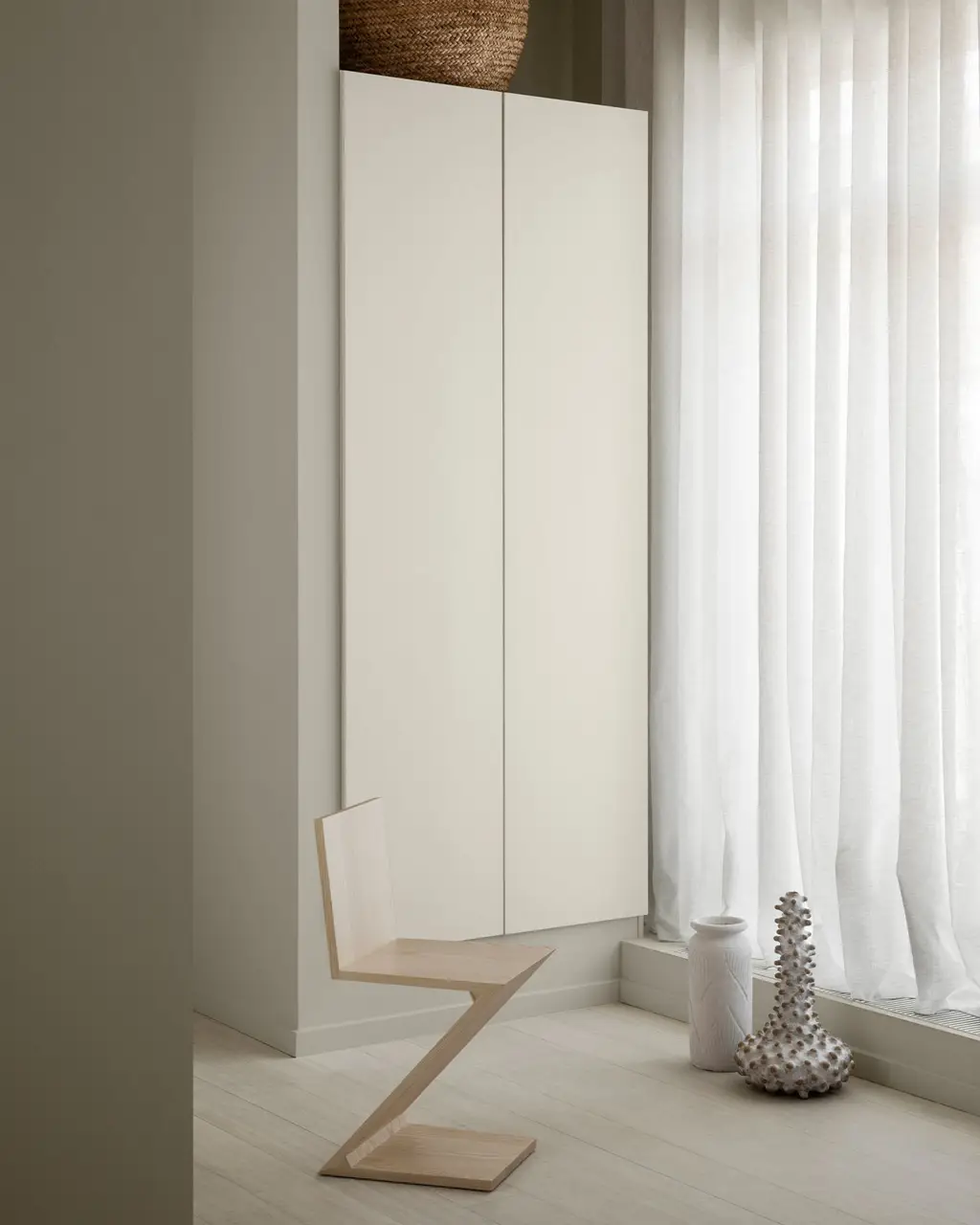Minimalistisk interiör med en geometrisk stol i trä, ett skåp målat i Jotun 1376 Froströk och tunna vita gardiner. Väggen framför är målad i 12180 Present, en dämpad beige nyans från Jotun.