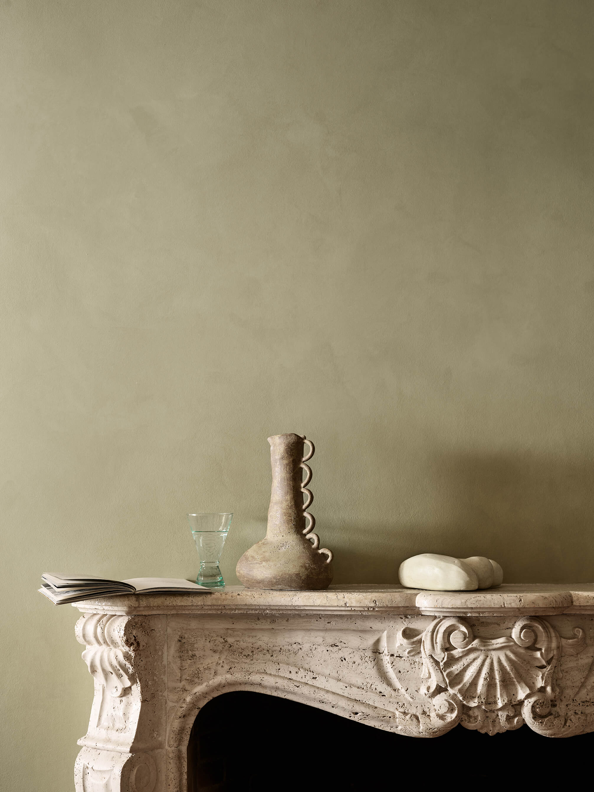 A refined living room featuring muted olive green walls in Jotun 0290 Biri Touch-of-Suede, an ornate stone fireplace mantel, and rustic ceramic decorative pieces