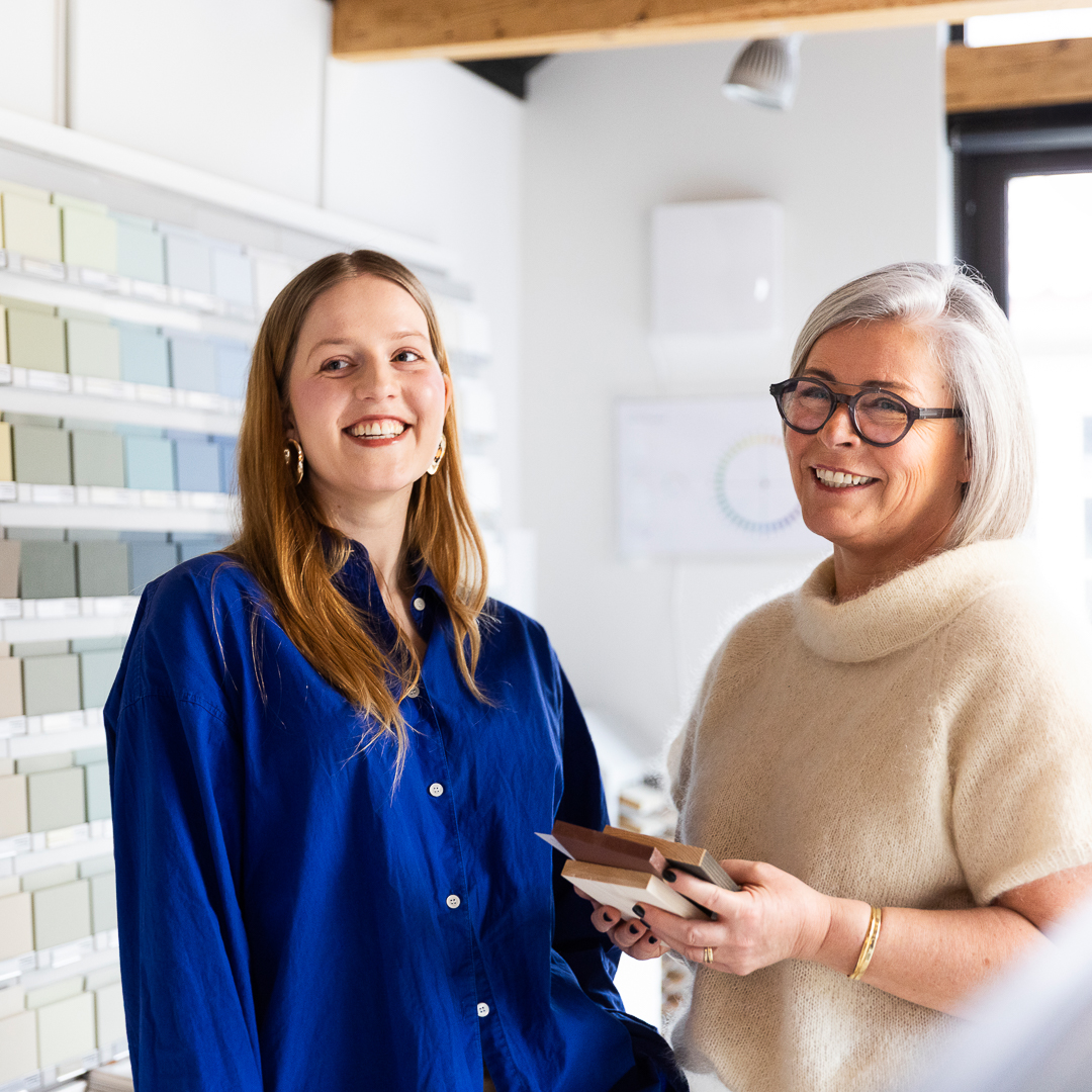 To fargerådgivere, smilende med en vegg med fargeprøver i bakgrunnen, en av dem holder tre fargeprøver fra Jotun i hånden