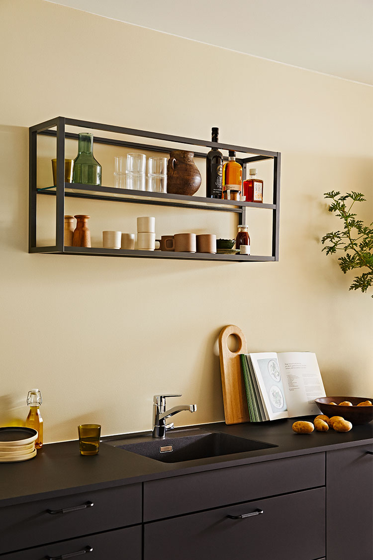 Moderne kjøkken med flytende hyller som fremviser glassvarer og flasker over mørk benkeplate med vask, skjærebrett og åpen kokebok, lysegul vegg i bakgrunnen malt i Jotun 12080 Soft Radiance