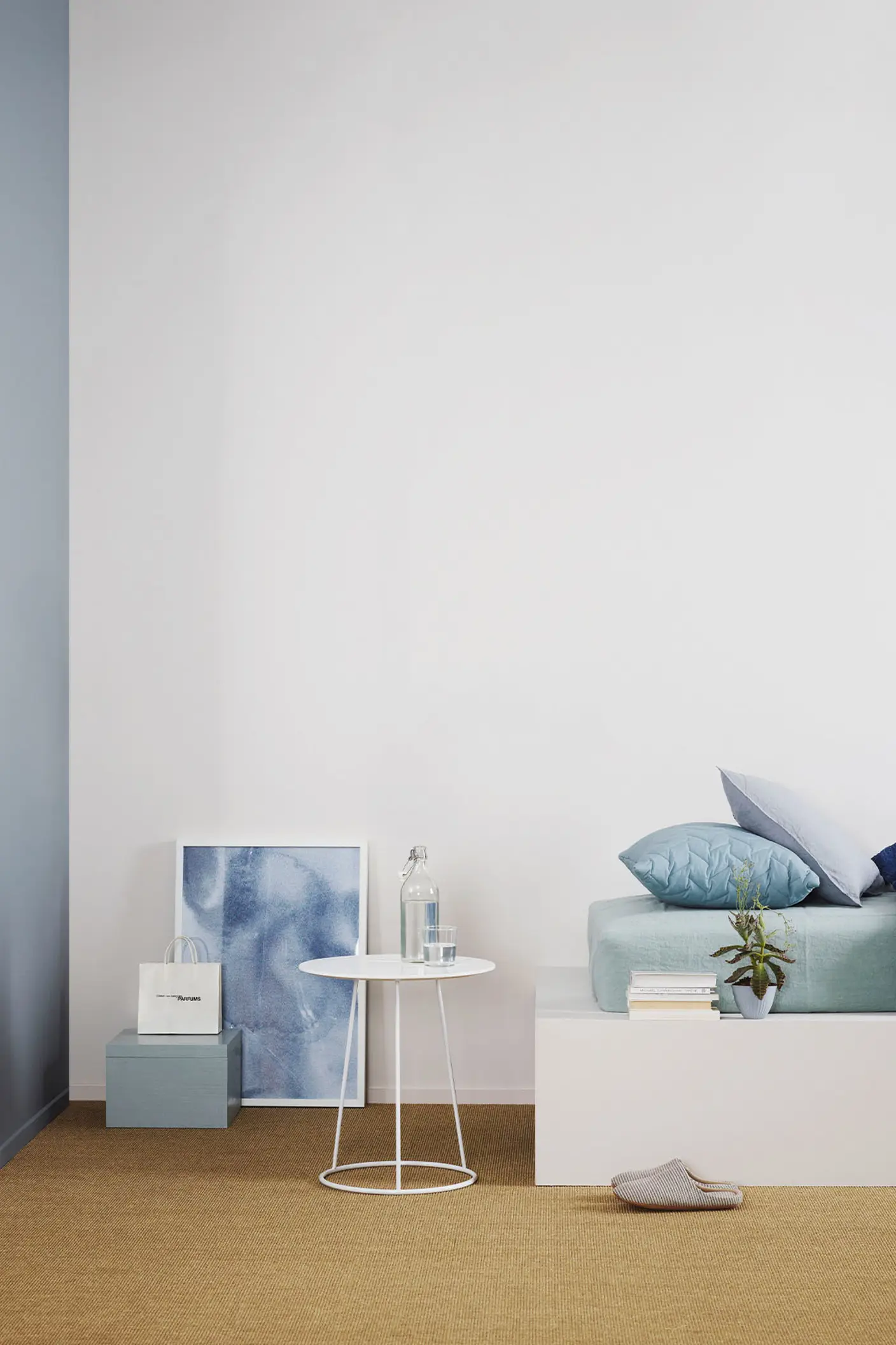 Minimalistiskt sovrum med en säng klädd i svala blå toner, ett vitt sidobord mot en gråvit vägg målad med Jotun 1622 Reflektion, och en dämpad rödblå accentvägg målad med Jotun 4710 Duggblå.