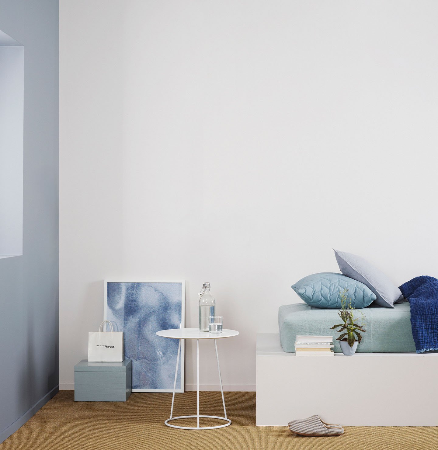 Minimalistiskt sovrum med en säng klädd i svala blå toner, ett vitt sidobord mot en gråvit vägg målad med Jotun 1622 Reflektion, och en dämpad rödblå accentvägg målad med Jotun 4710 Duggblå.