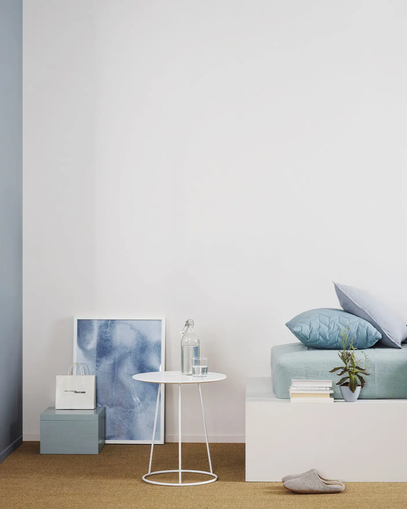 Minimalistiskt sovrum med en säng klädd i svala blå toner, ett vitt sidobord mot en gråvit vägg målad med Jotun 1622 Reflektion, och en dämpad rödblå accentvägg målad med Jotun 4710 Duggblå.