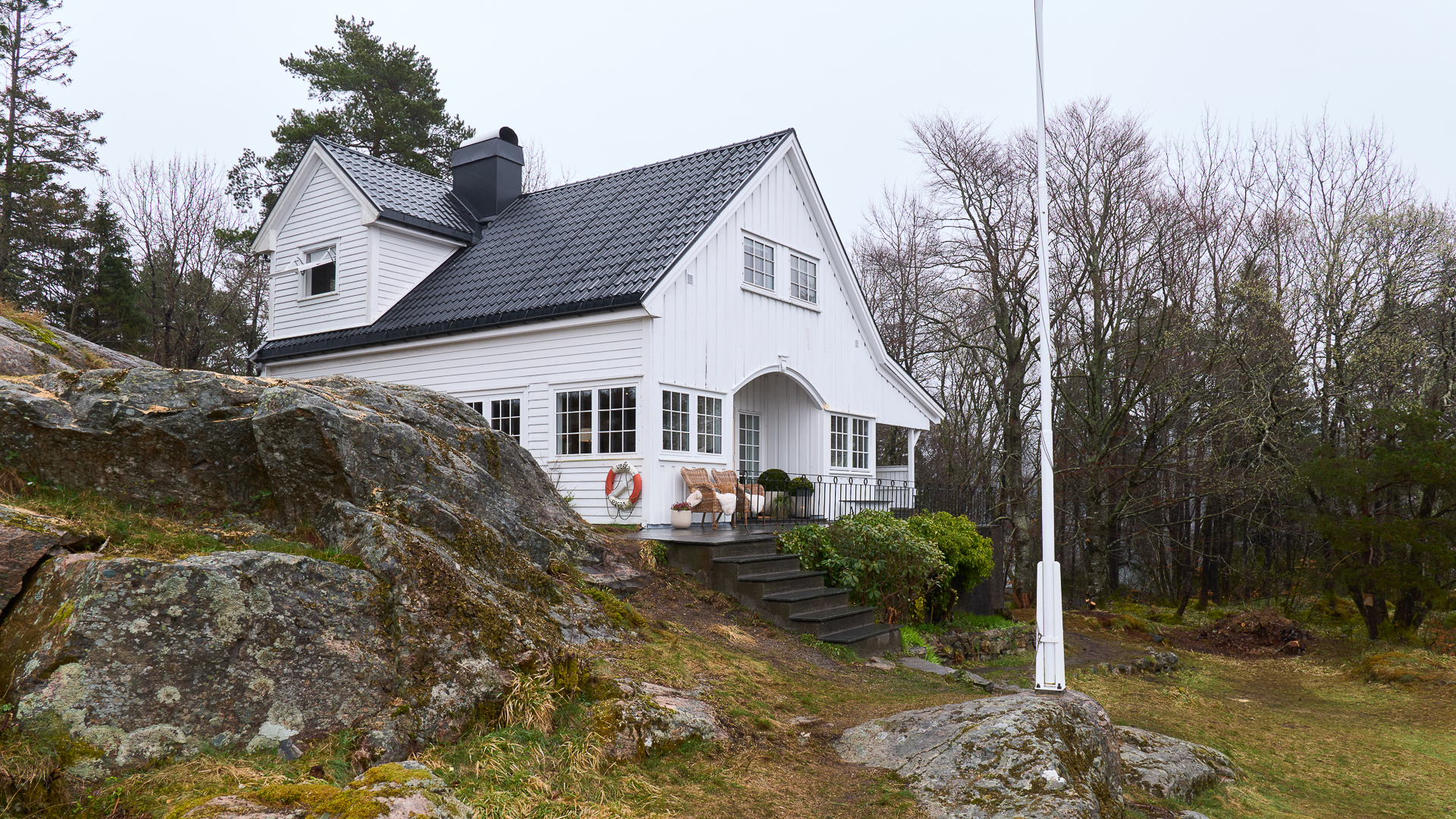 Hvit trehytte med svart tak i landlig skogsområde, omgitt av steiner og trær