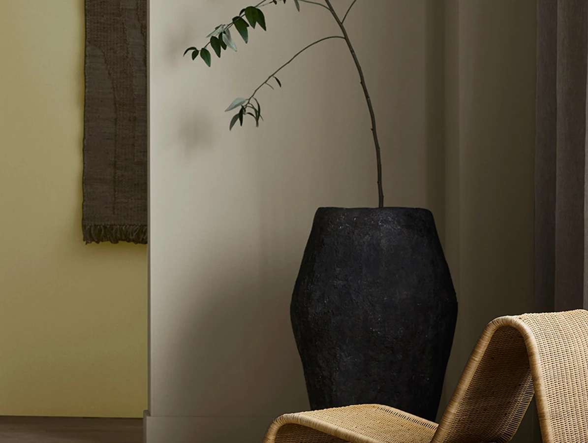 Woven lounge chair next to a tall black vase with a branch, in front of a doorway opening to a muted yellow room