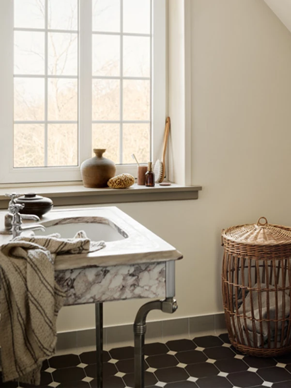 Moderne bad med malte vegger i beige i fargen Jotun 12079 Gleam, marmorbaderomsvask, stripete håndkle og et vindu som slipper inn naturlig lys. Vase og toalettartikler på vinduskarmen, samt en flettet skittentøyskurv på gulvet med sekskantede fliser i sort og hvitt
