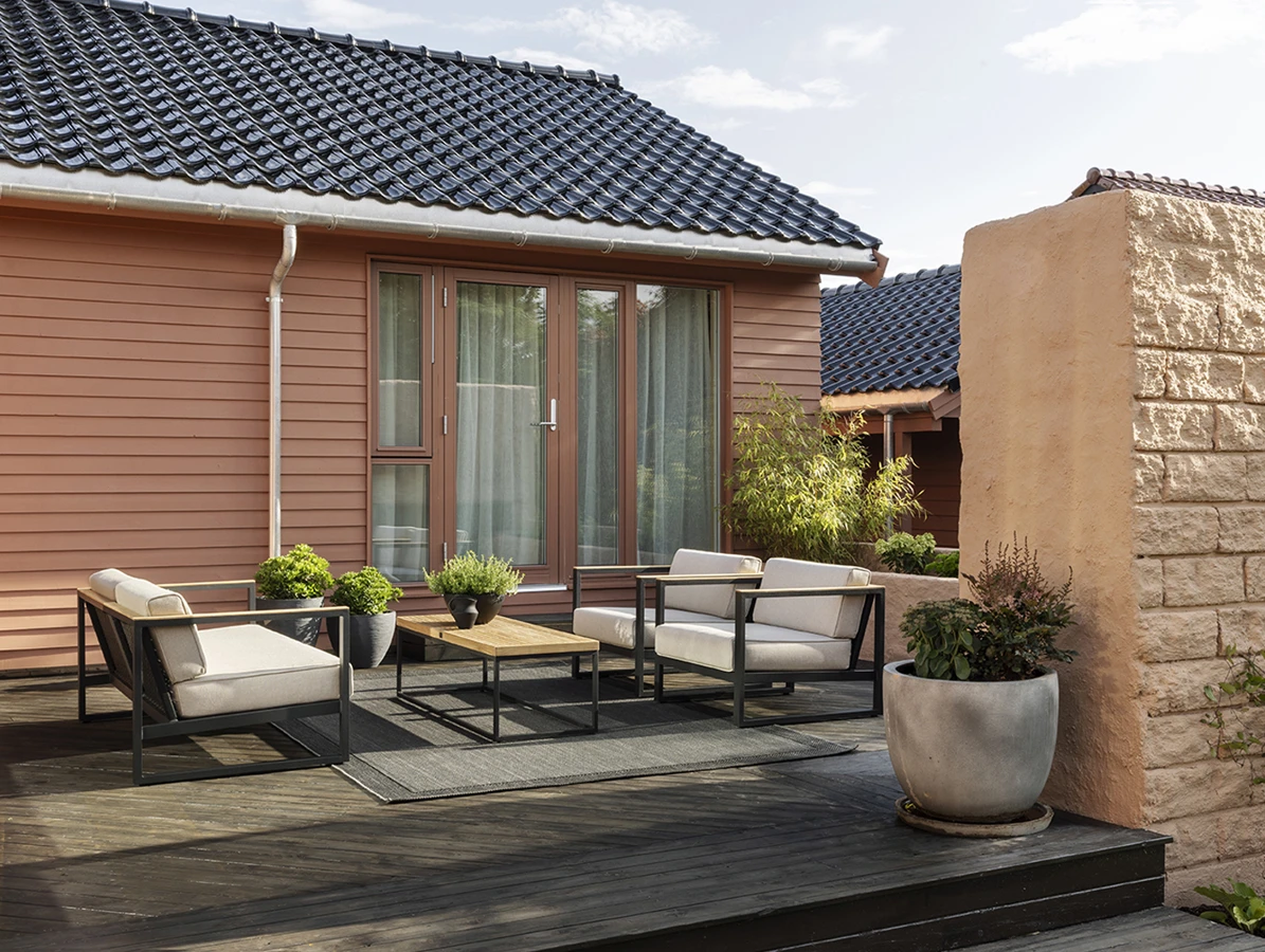 Modern outdoor patio with black metal chairs and beige cushions on a wooden deck, adjacent to a house with red siding and large windows, surrounded by potted plants and a stone wall