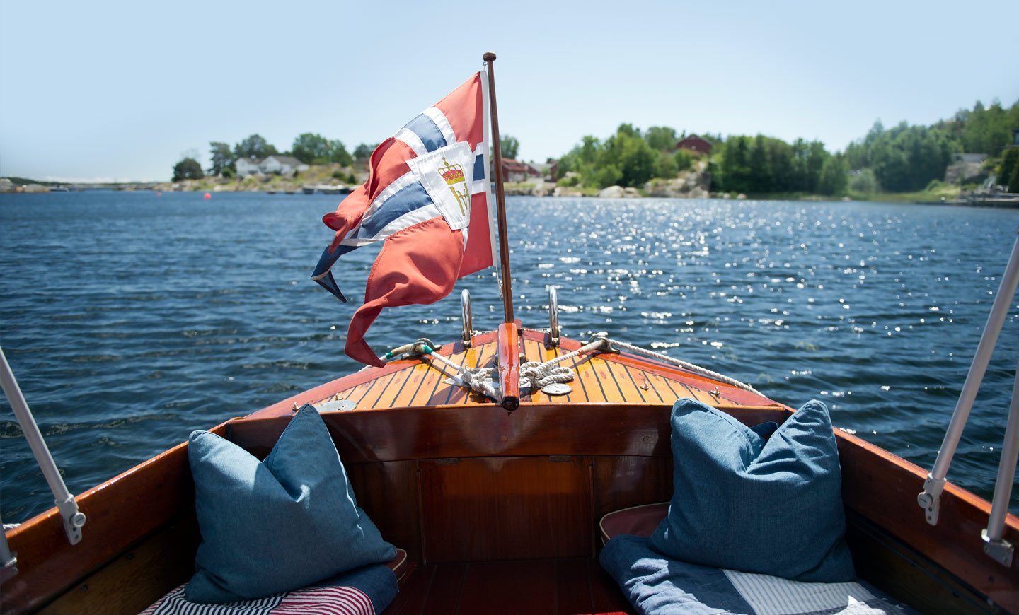 Akterdekk på trebåt med norsk flagg som ligger på rolig vann i sommersol.