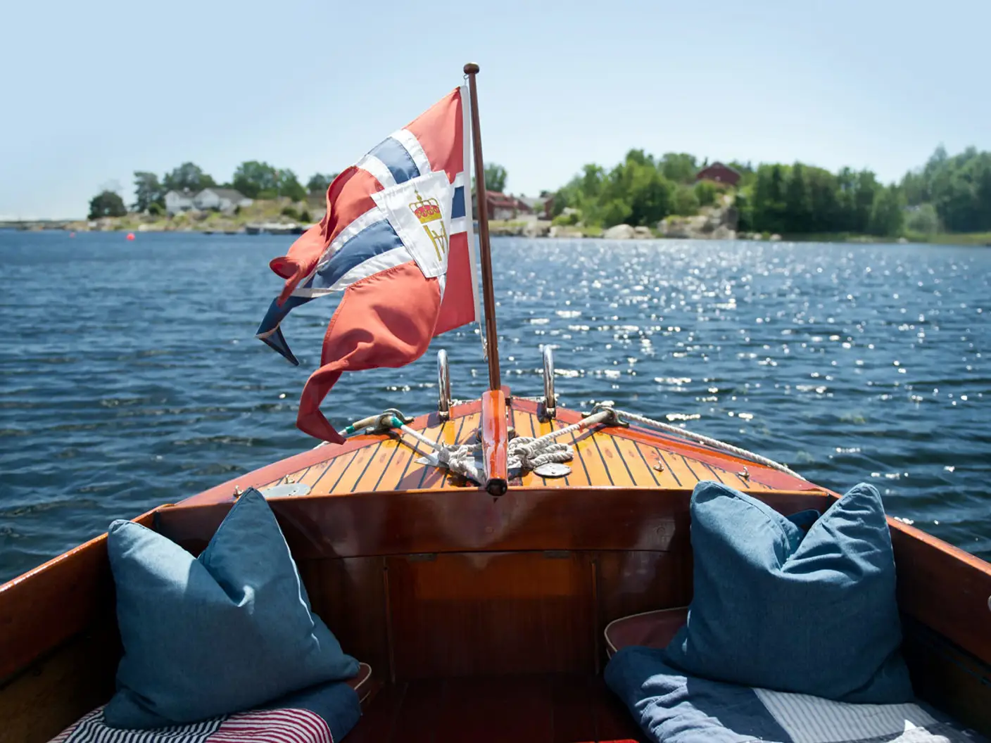 Akterdekk på trebåt med norsk flagg som ligger på rolig vann i sommersol.