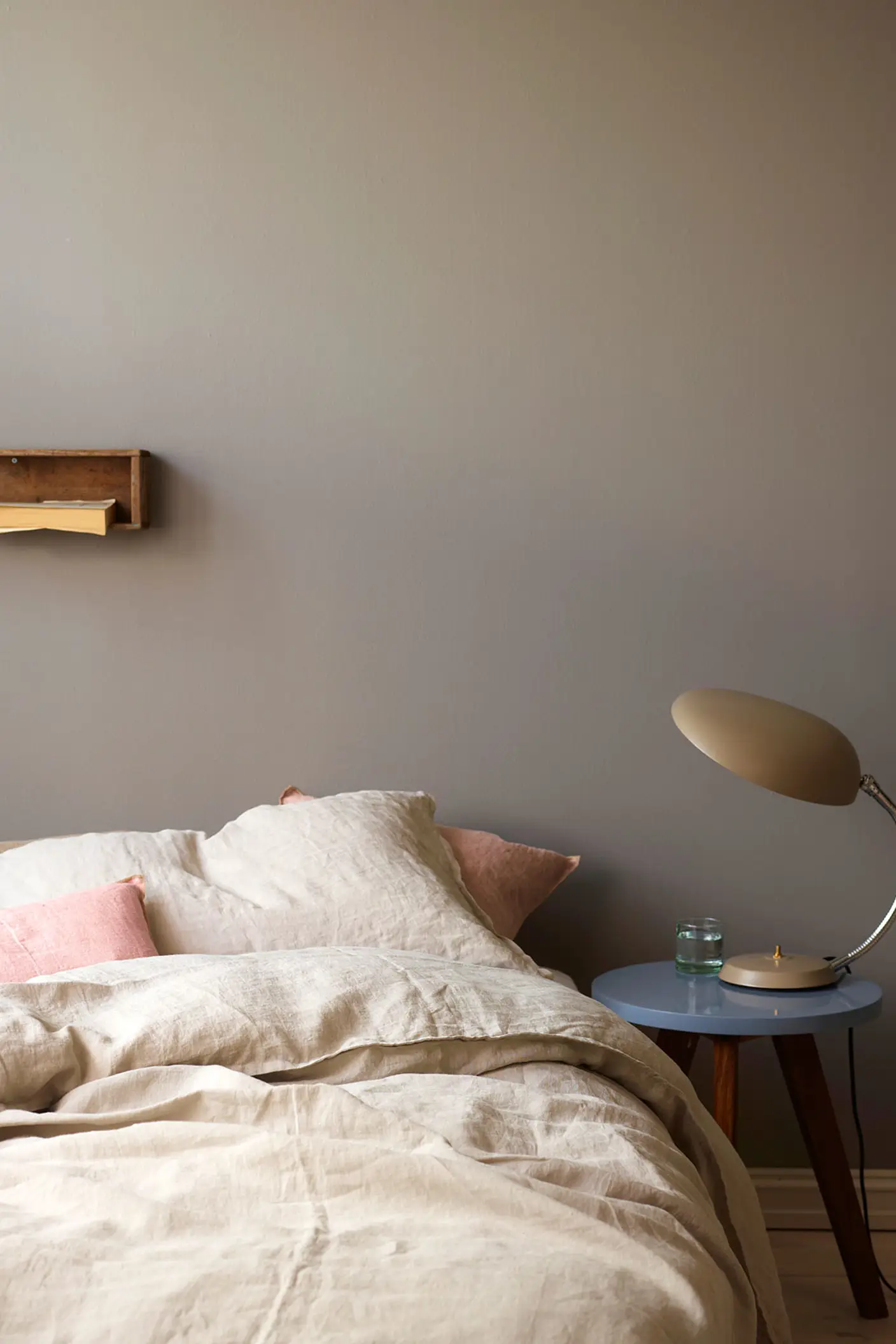 Nötr tonlu keten nevresimli ve açık pembe yastıklı yatak; retro lambalı yan sehpa ile birlikte, Jotun 1877 Pebblestone tonuyla boyanmış altın gri bir duvarın önünde yer alıyor.