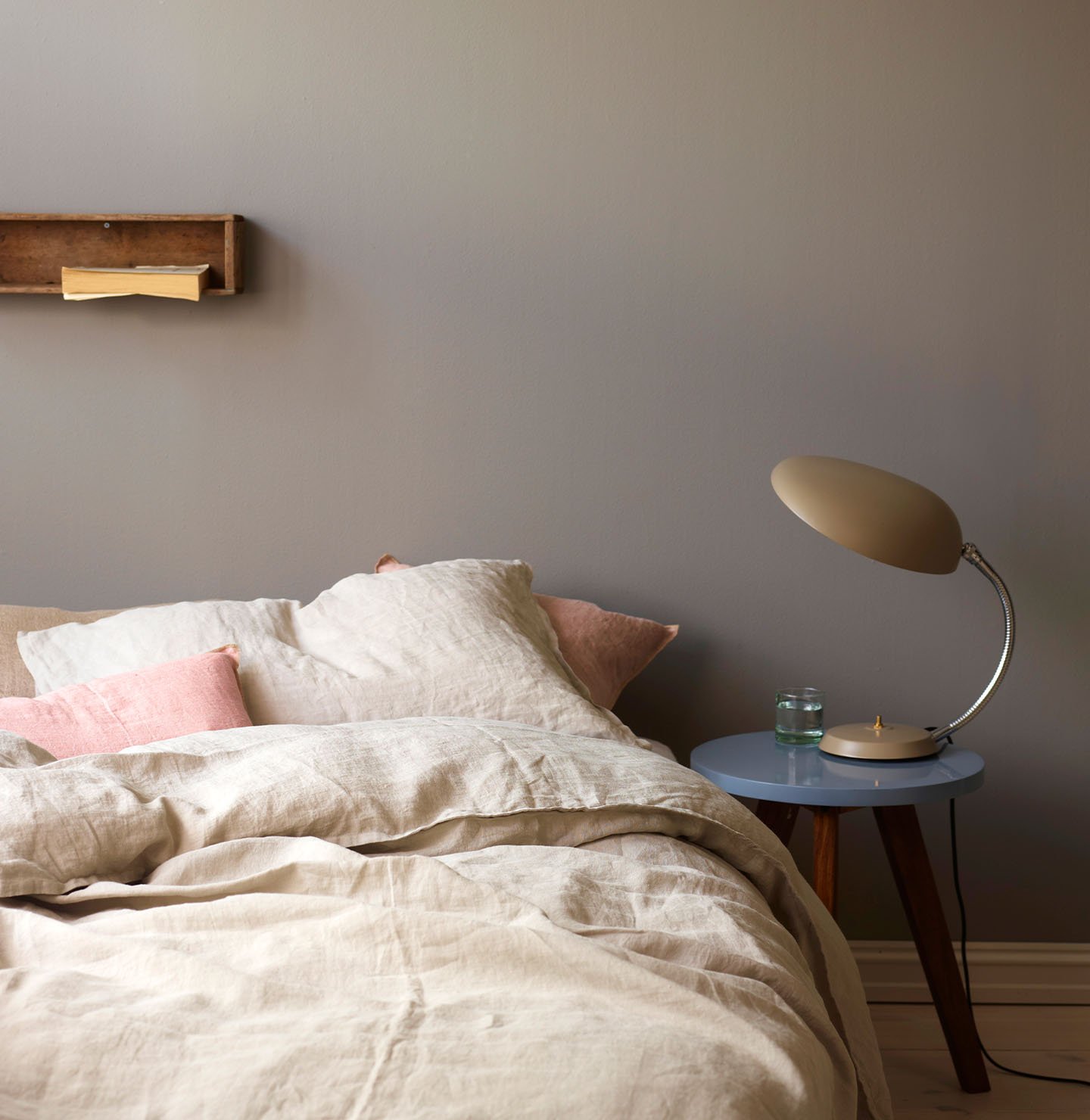 Neutral-toned bed with soft linen bedding and pale pink accent pillows, beside a  side table holding a retro lamp, against a golden grey wall painted with Jotun 1877 Pebblestone.