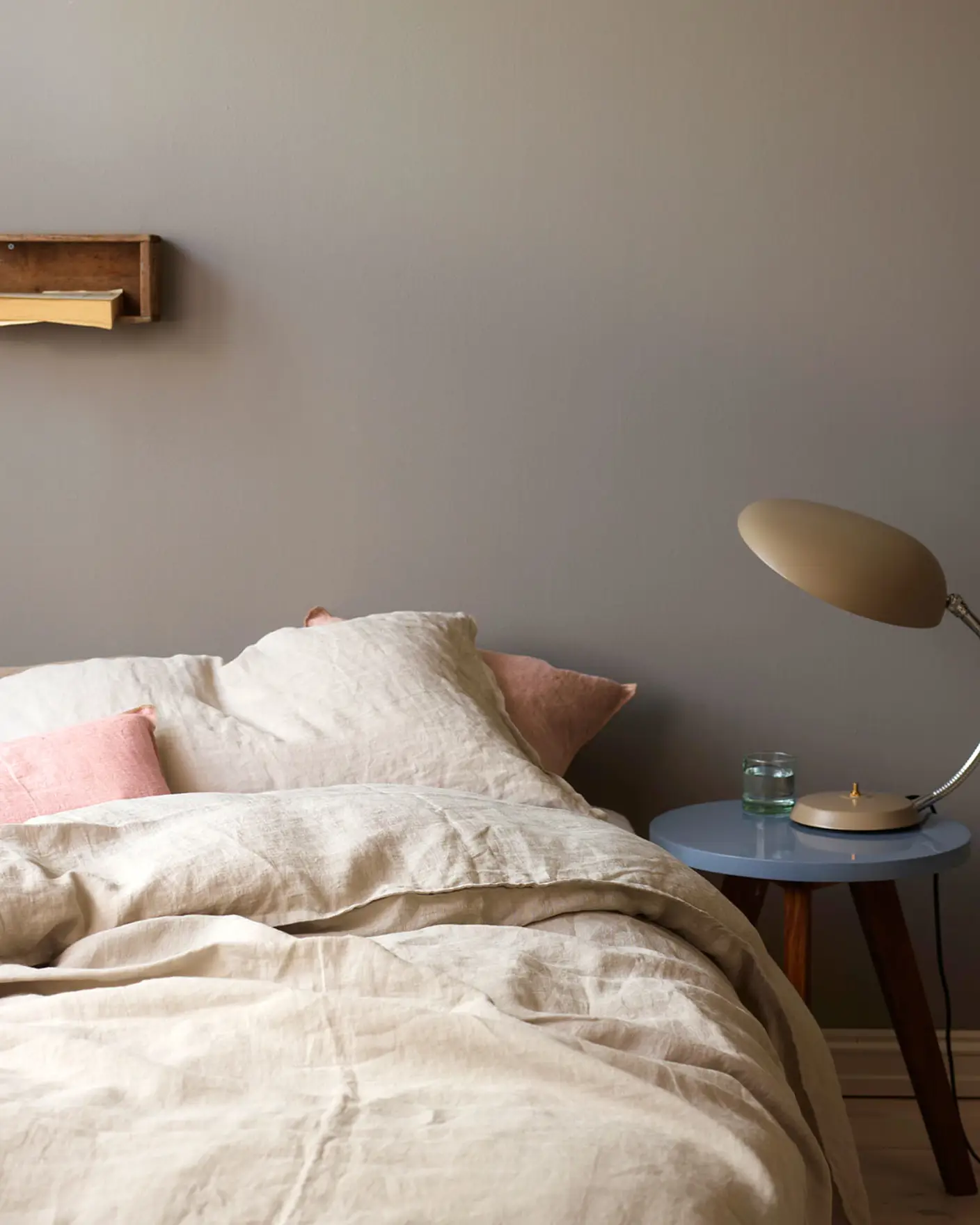 Neutral-toned bed with soft linen bedding and pale pink accent pillows, beside a  side table holding a retro lamp, against a golden grey wall painted with Jotun 1877 Pebblestone.