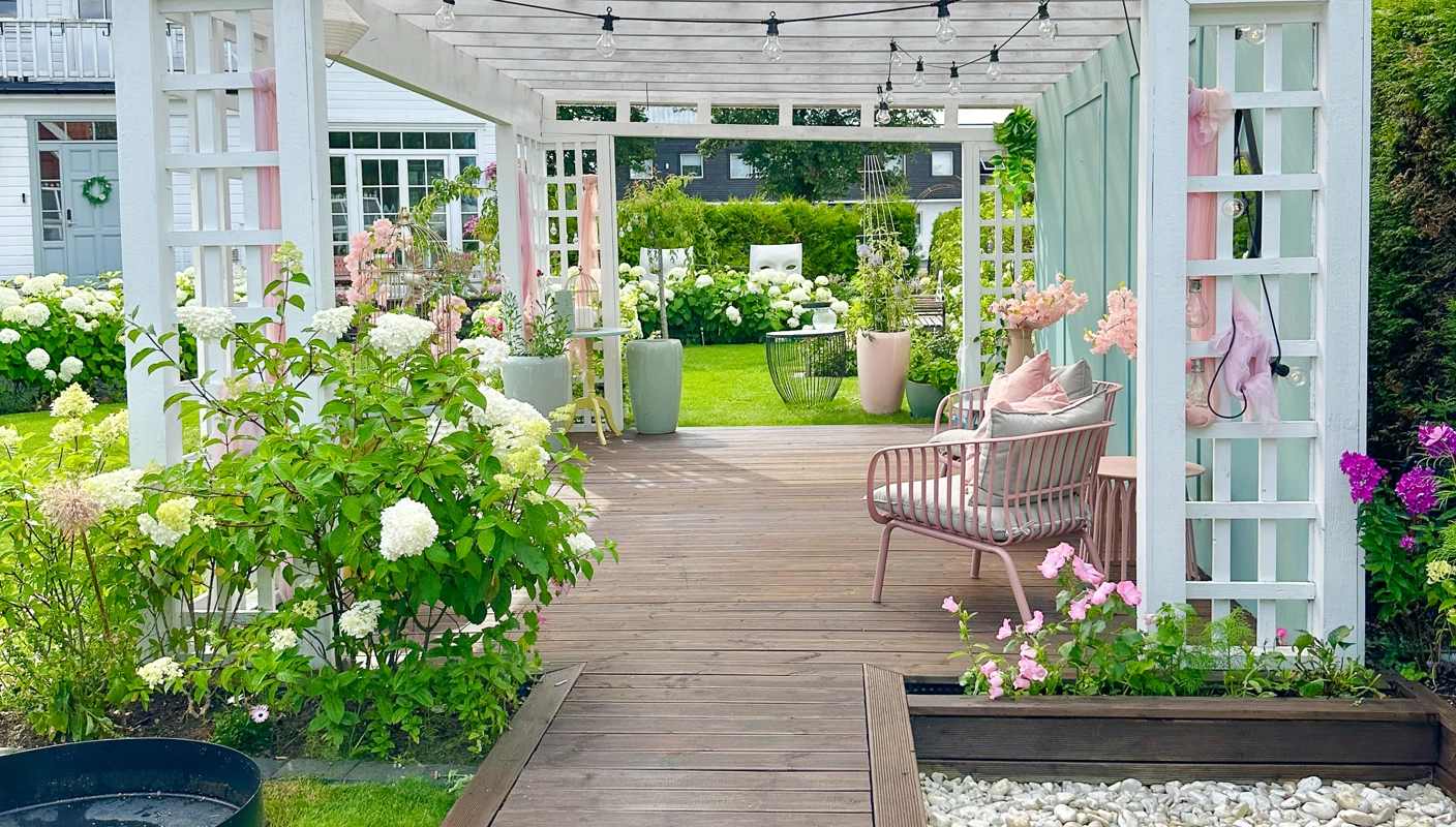 Brunt terrassegulv beiset med TREBITT Terrassebeis i Terrassebrun, ute i en grønn hage med en pergola malt hvit med JOTUN Klassisk Hvit