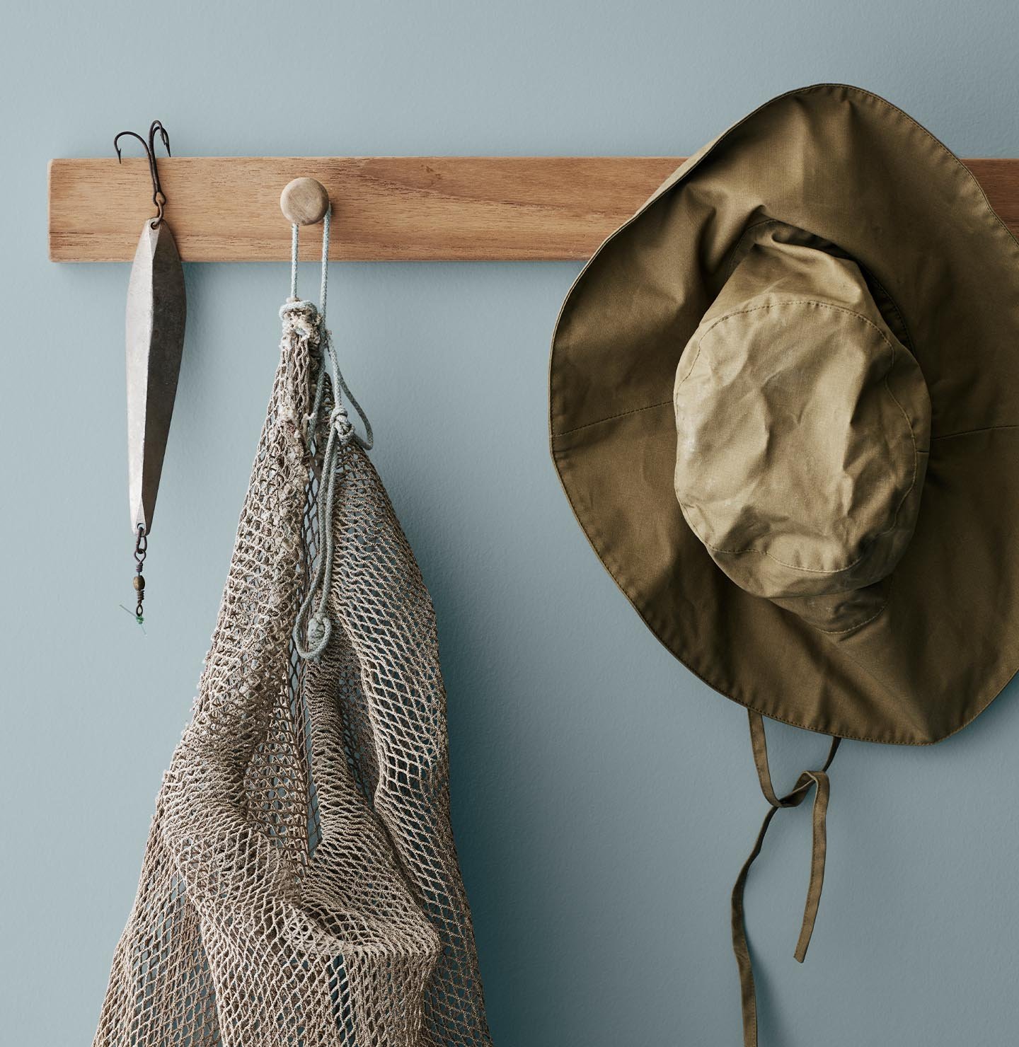 Et trehyllestativ holder et fiskeagn, et nett og en olivengrønn hatt, mot en dempet blå vegg, malt med Jotun 4894 Ocean Air, som gir en luftig kystfølelse.