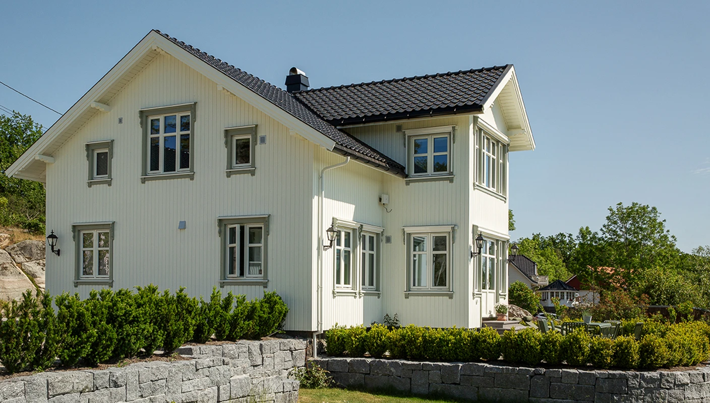 Hvitt klassisk trehus malt hvitt i Jotun Bomull med DRYGOLIN Power Clean, sort takstein og  grønn hekk runt huset.