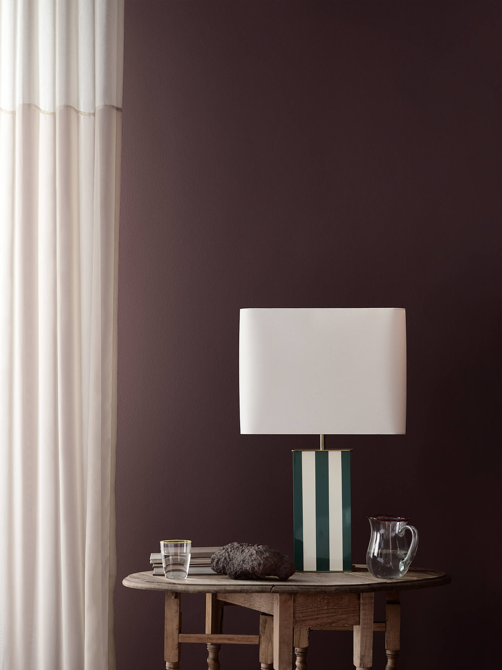 I et hjørne av stuen er veggen malt i den mørke lillafargen Jotun 1090 Dusty Purple, og her står en stripete bordlampe på et rustikt trebord, samt hvite, gjennomsiktige gardiner.