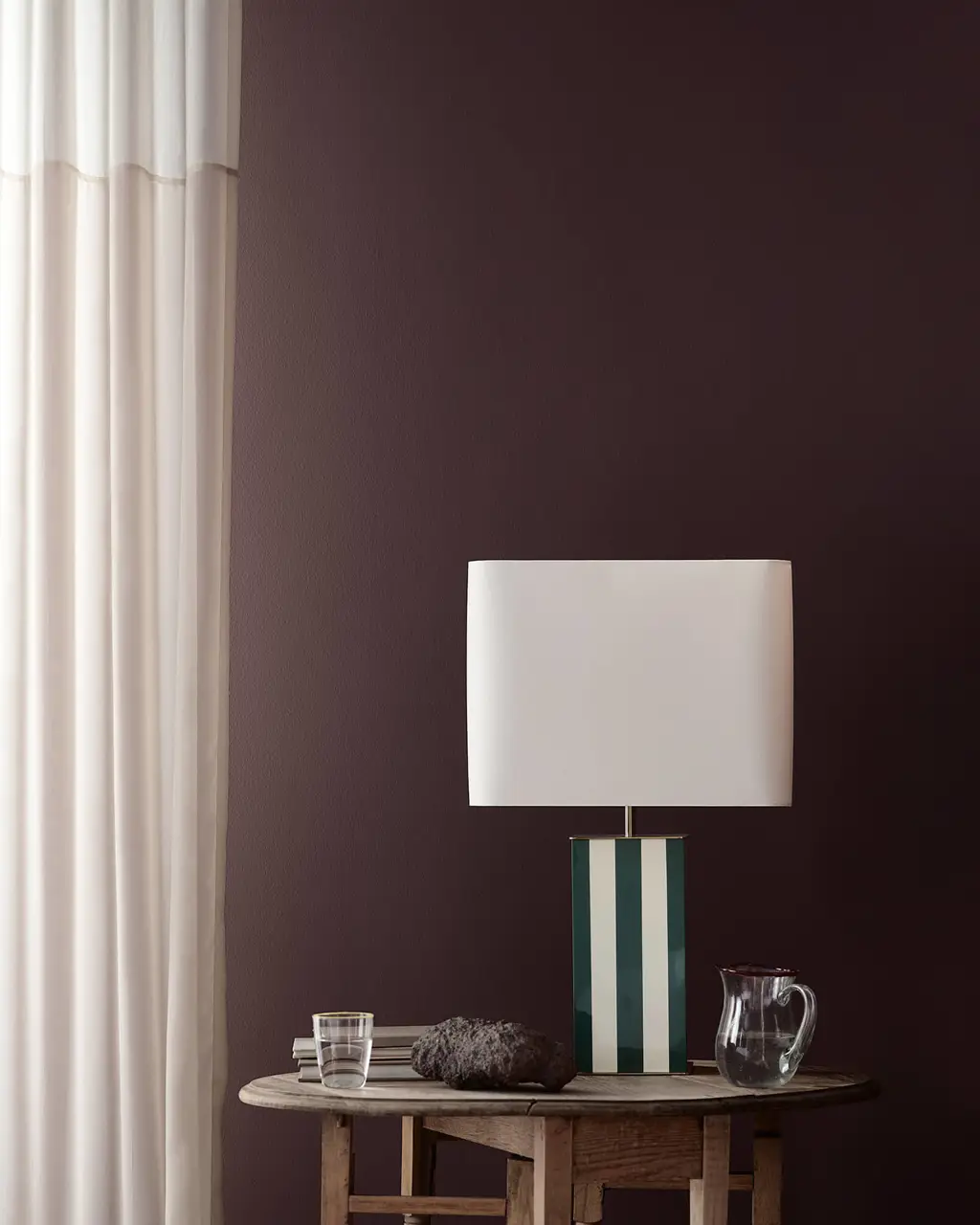 I et hjørne av stuen er veggen malt i den mørke lillafargen Jotun 1090 Dusty Purple, og her står en stripete bordlampe på et rustikt trebord, samt hvite, gjennomsiktige gardiner.