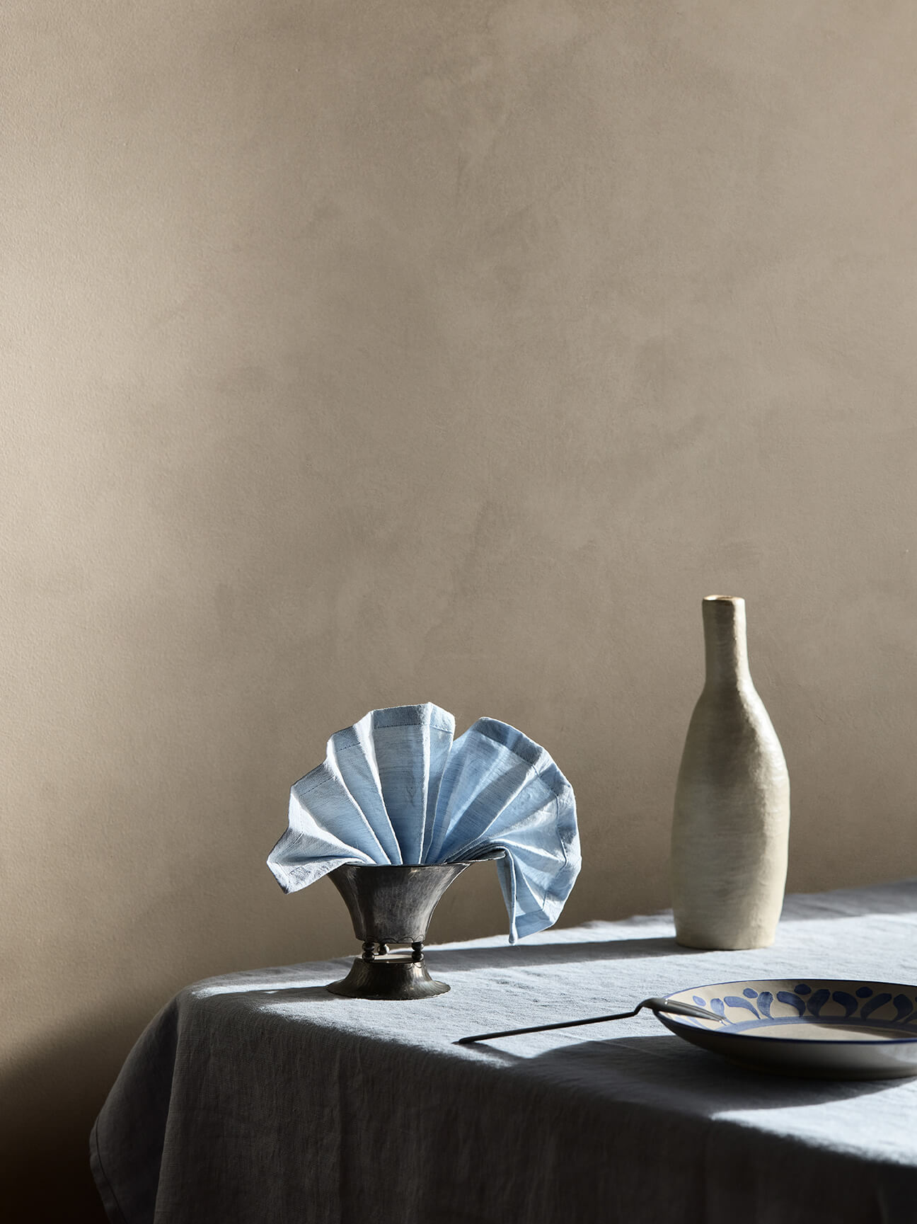 A close-up of a table setting with a blue napkin and ceramic vase against a textured wall painted in Jotun 10966 Almond Beige Touch of Suede