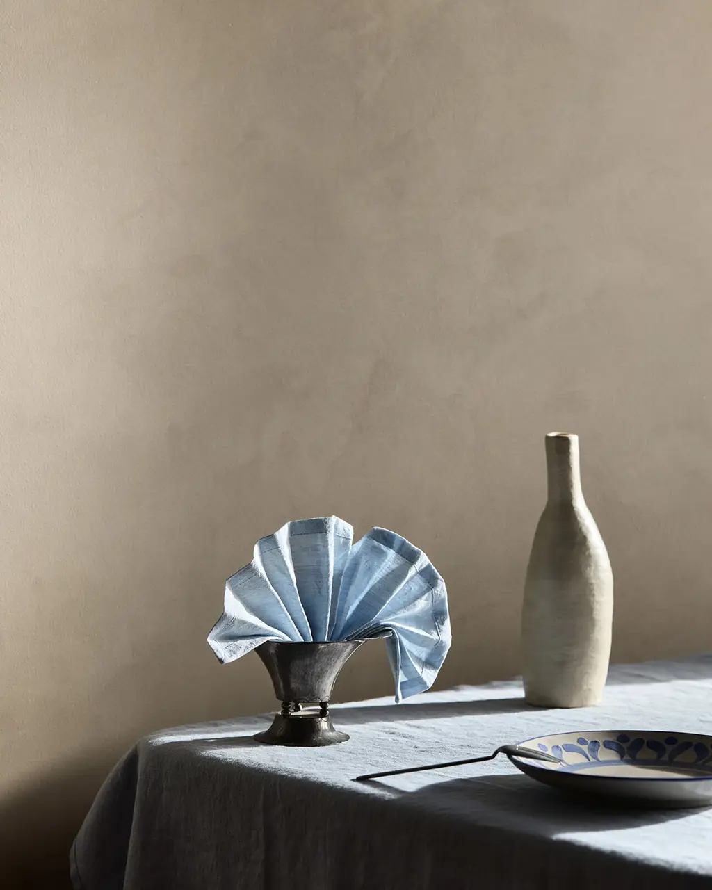 A close-up of a table setting with a blue napkin and ceramic vase against a textured wall painted in Jotun 10966 Almond Beige Touch of Suede