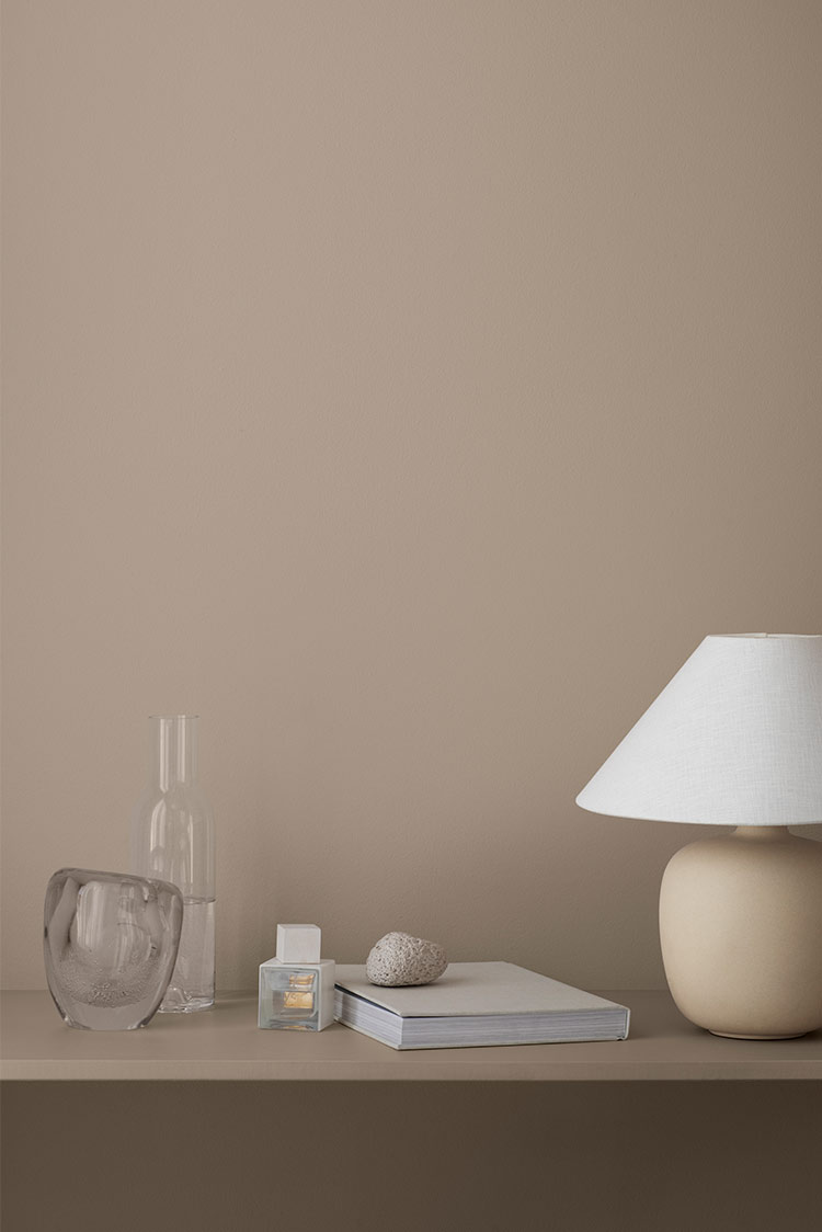Minimalistisk interiør med beige vegg malt i Jotun 20184 Thoughtful, dekorativ bordlampe, glasskarafler, bøker og en liten stein på en hylle. Naturlige jordtoner skaper en rolig atmosfære