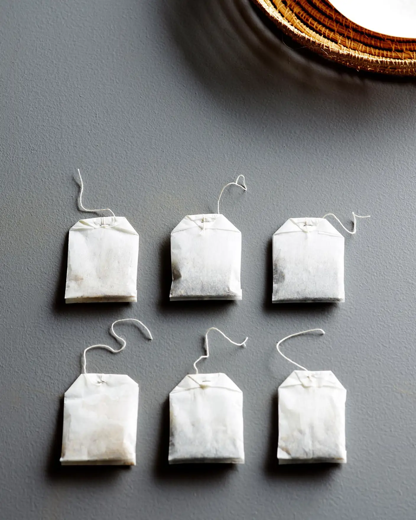 Tepåsar ligger prydligt arrangerade på en texturerad blågrå yta målad med färgen Jotun 4629 Skuggblå, bredvid en vävd tallrik, vilket skapar en lugn och minimalistisk känsla.