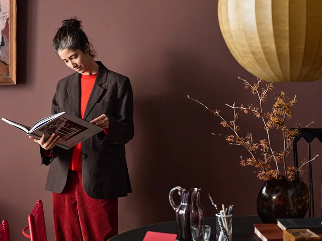 Person som läser en bok i ett rum med mörka väggar målad i Jotun 2951 Sophisticated Red, med moderna möbler, ett runt svart bord och en stor hängande lampa