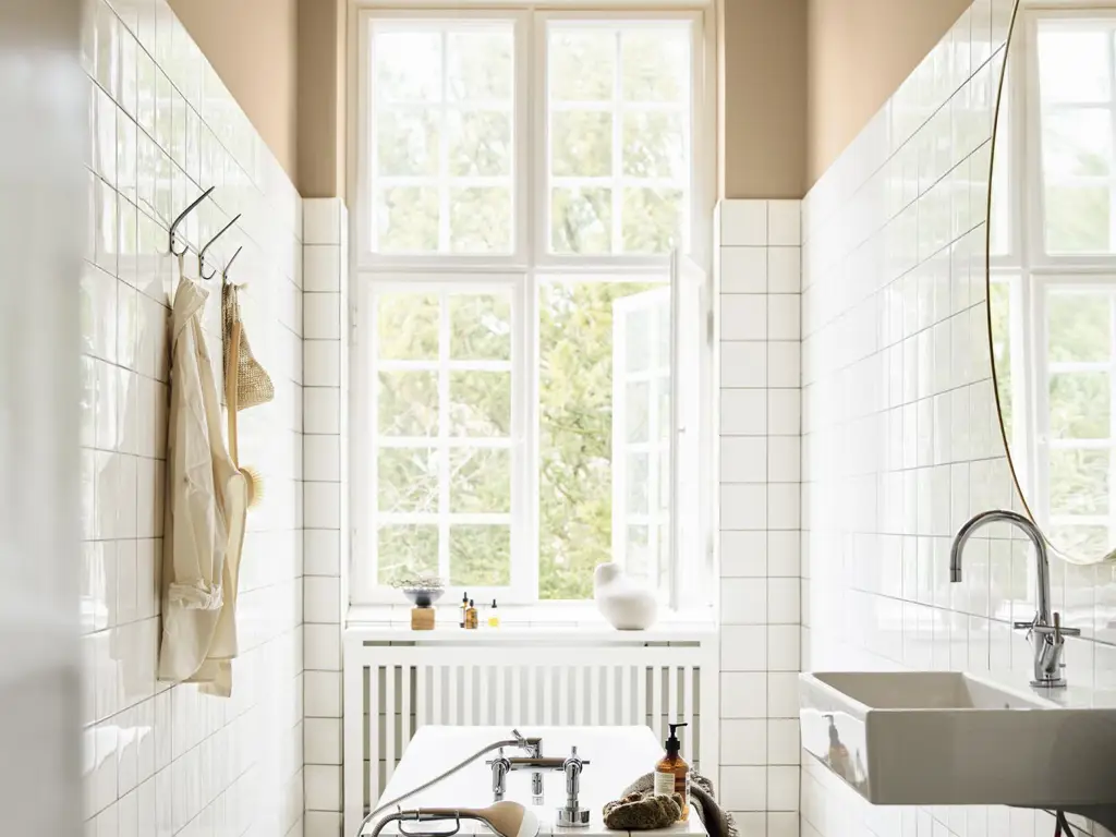Lyst og minimalistisk baderom med store vinduer, hvite fliser, frittstående badekar og servant med speil.