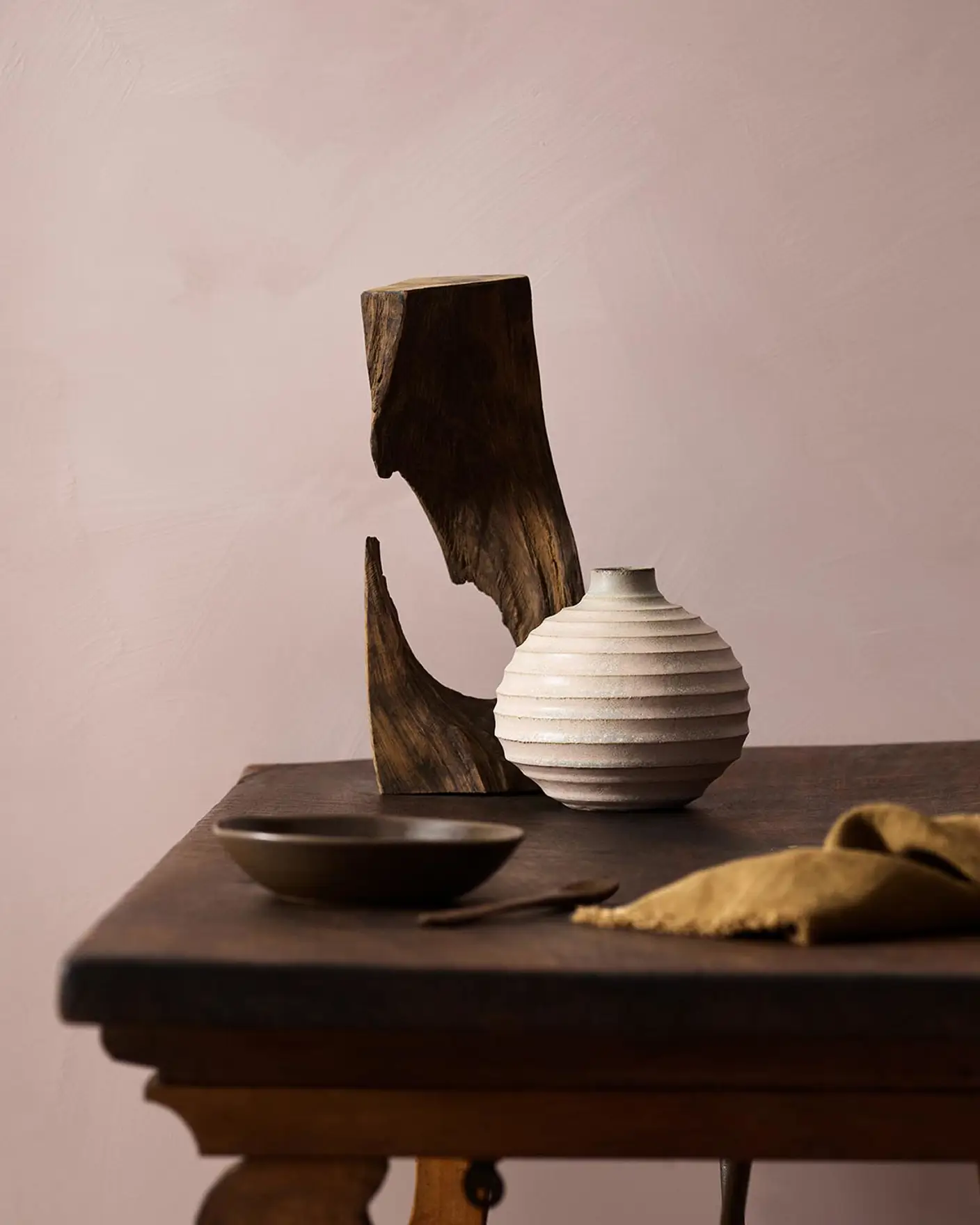 Wooden table with a ceramic vase, sculpted wood, and a dark bowl against a muted pink background painted with LADY Design Touch of Suede in the colour 20054 Silky Pink from Jotun.