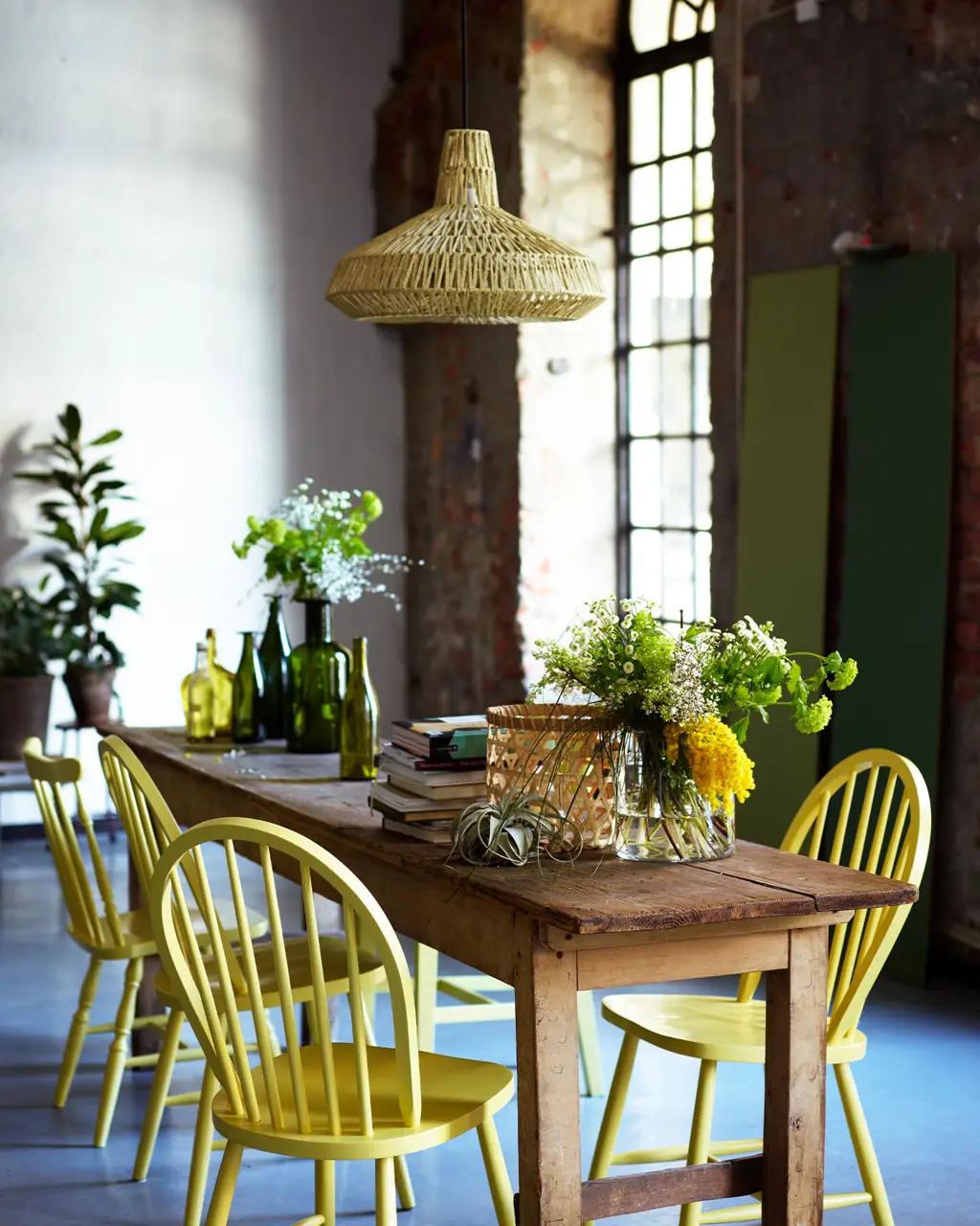 Un espacio de comedor rústico con sillas pintadas de amarillo fresco, en el color 10235 Summer Sun de Jotun, rodeadas de suave verde y texturas industriales.