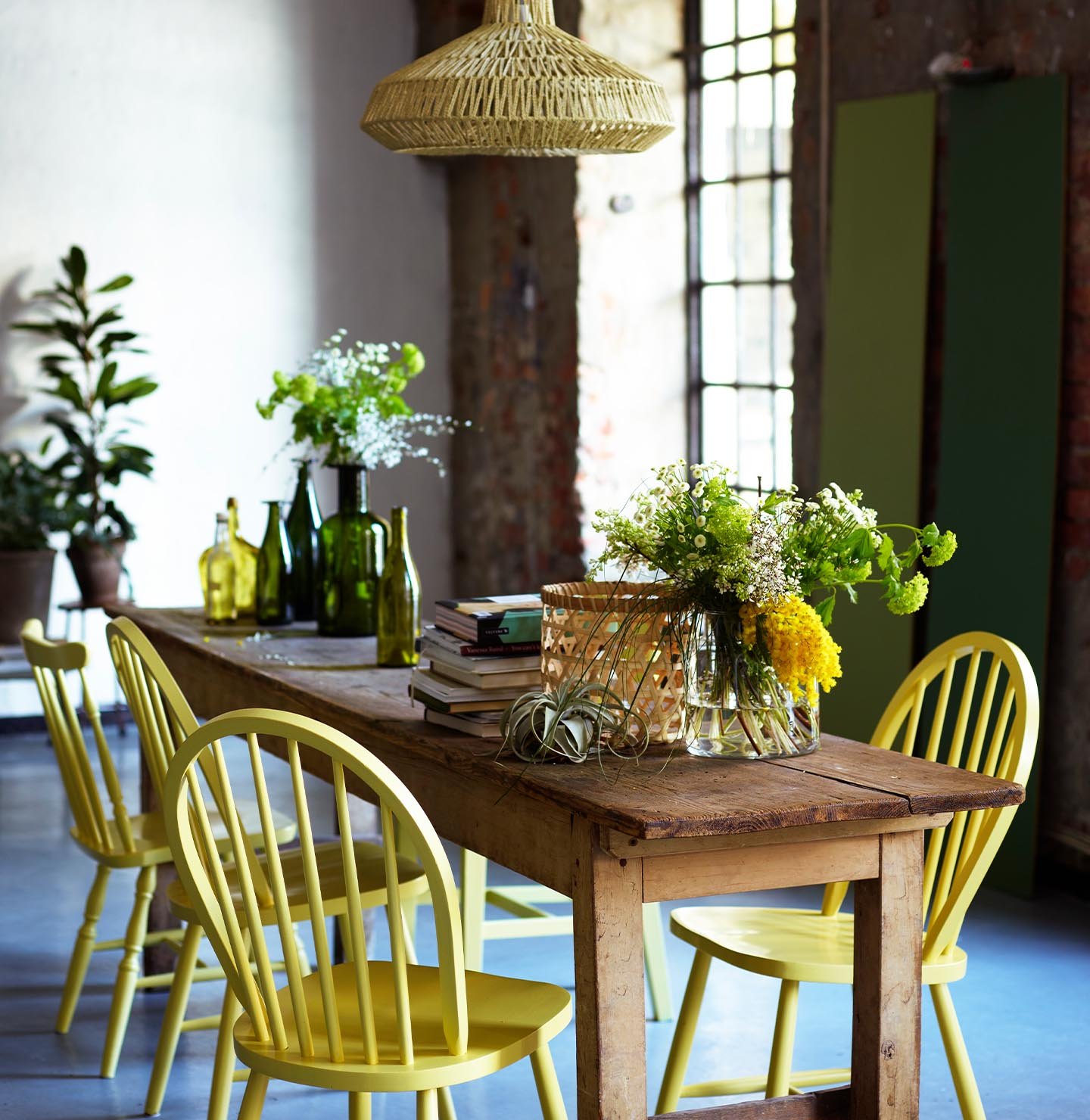 Rustik matplats med stolar målade i en frisk gul nyans, färgen 10235 Summarsol från Jotun, omgiven av mjuka gröna toner och industriella texturer.