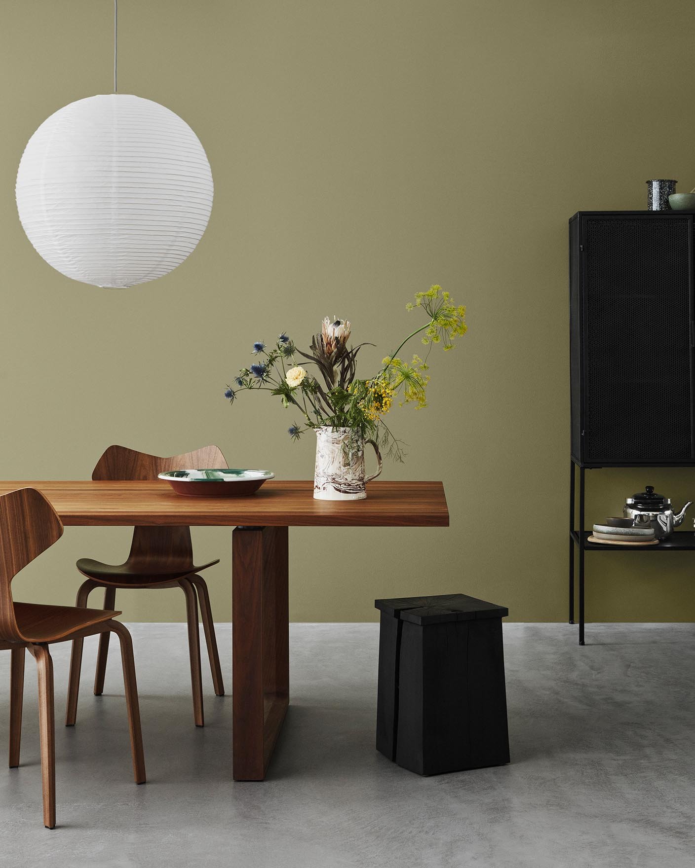 A minimalist dining space with wooden furniture set against an olive green wall, painted with Jotun 8546 Local Green, and concrete floor.