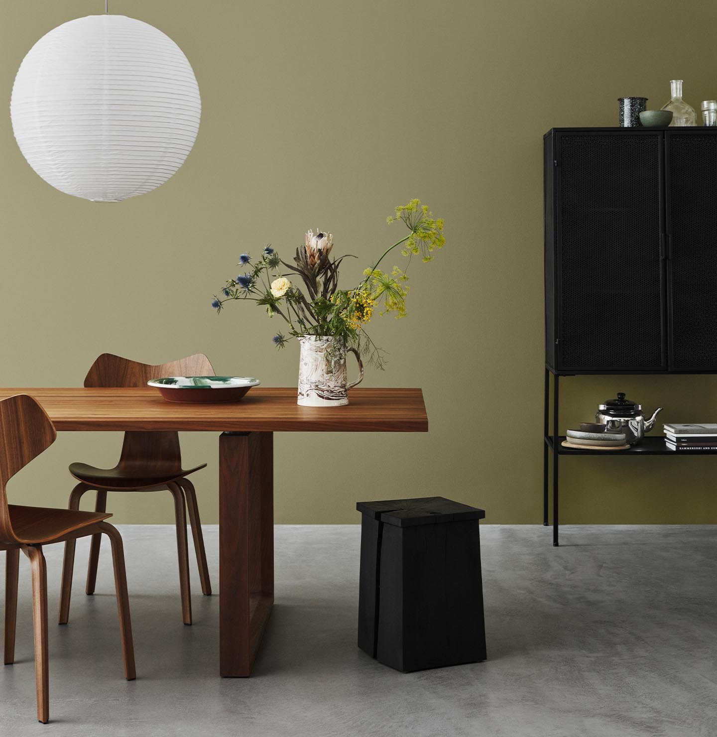 A minimalist dining space with wooden furniture set against an olive green wall, painted with Jotun 8546 Local Green, and concrete floor.