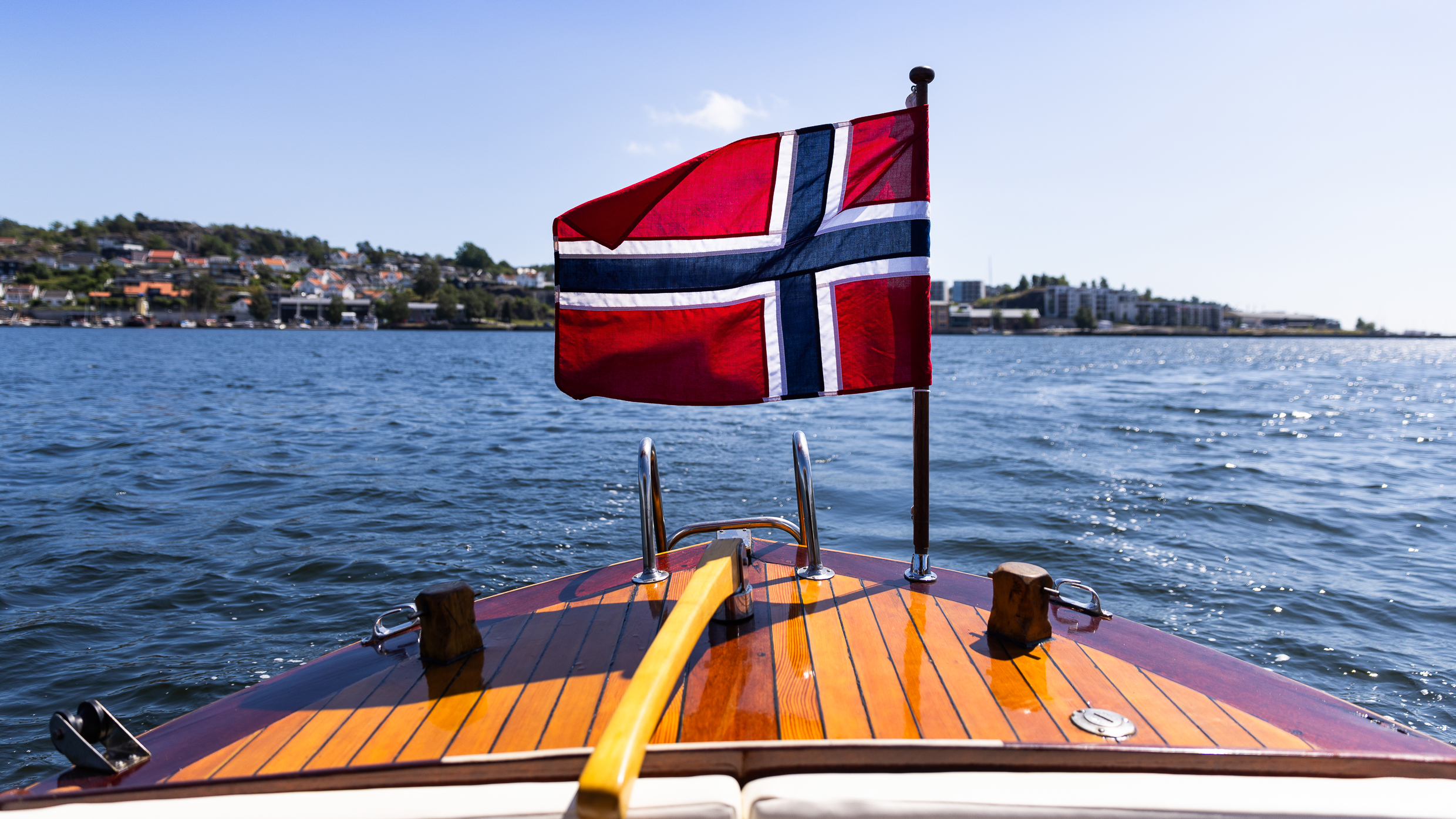 Norsk flagg på blankpusset trebåt omringet av blå himmel, blå fjord og sollys