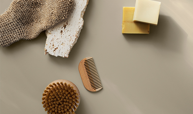 Naturlige badeprodukter på greige overflate: strikket vaskevott, to såpestykker, trekam, skrubbebørste og en rustikk flisstein