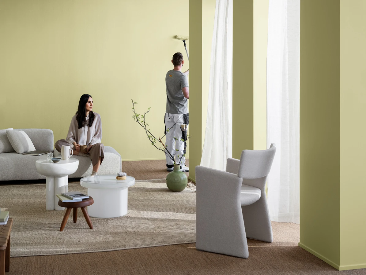 Açık renkli modern oturma odasında duvar boyayan bir kişi, kanepede oturan bir kadın ve minimalist dekorlu salon düzeni