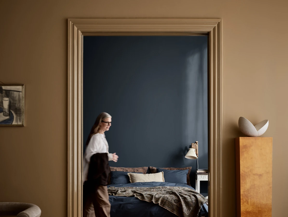 Soverom sett gjennom en stor døråpning, med mørk blå vegg i fargen Midnight Blue fra Jotun, oppredd seng og en kvinne som går forbi.
