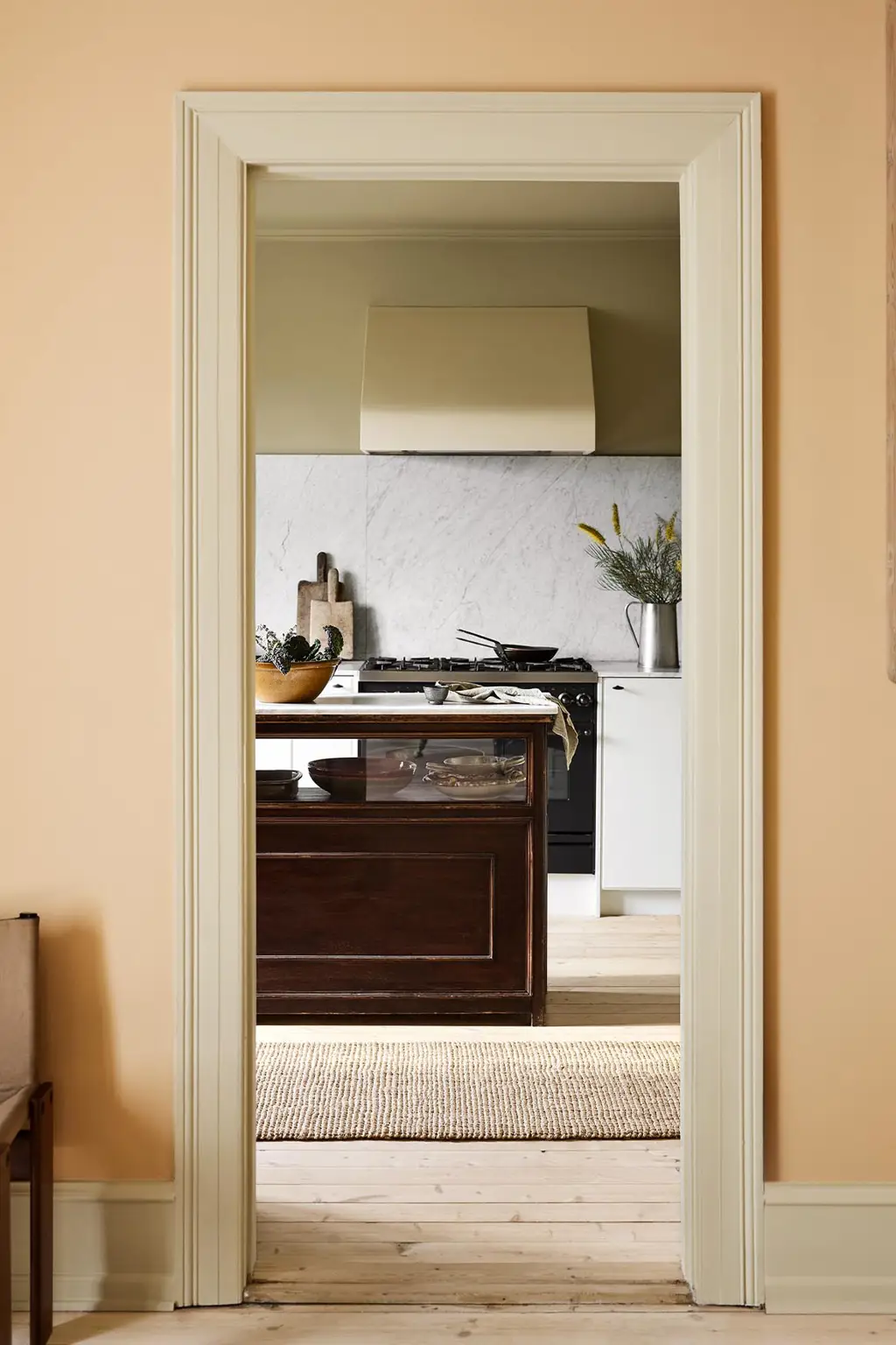 Kitchen viewed through a doorway. The wall in front is painted in the pale, ochre yellow colour 11202 Mild Ochre from Jotun.