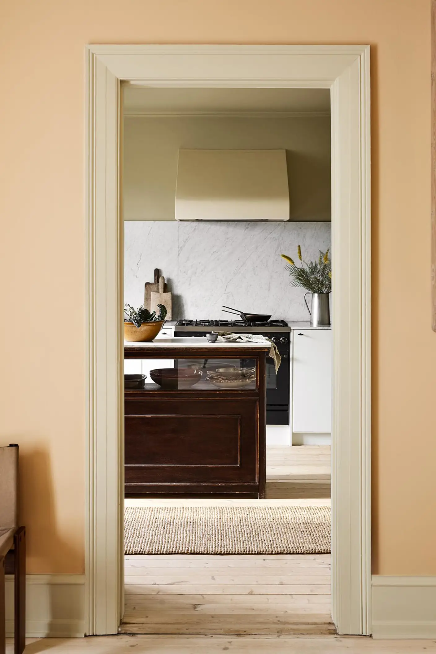 Kitchen viewed through a doorway. The wall in front is painted in the pale, ochre yellow colour 11202 Mild Ochre from Jotun.