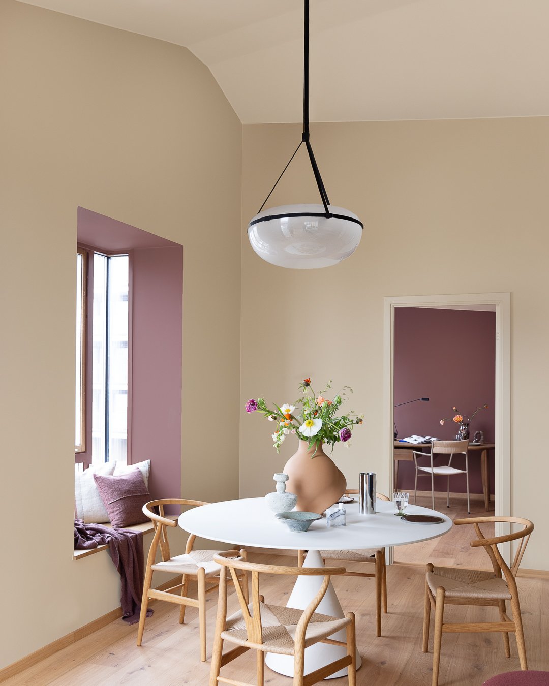 Dinigroom with modern chairs in oak, a round table with organically shaped vase in terracotta with colourful flowers, beige wall in the background painted in JOTUN 1303 Observe, with a deep red accent colour in the window frame painted in JOTUN 2951 Sophisticated Red.