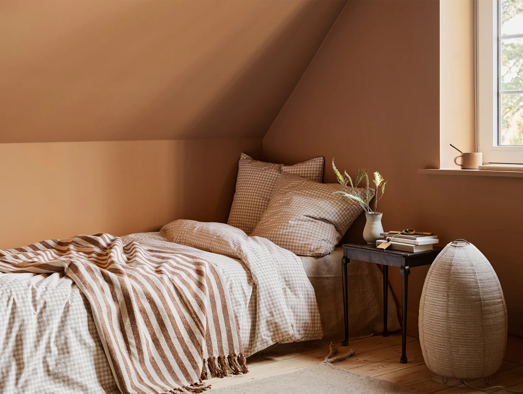 Koselig soverom malt i varm okergul med Jotun 11220 Ochre Clay, med seng dekket av stripete og rutete sengetøy, plassert ved vindu og småbord med dekorativ vase og bøker, i et varmt og behagelig interiør